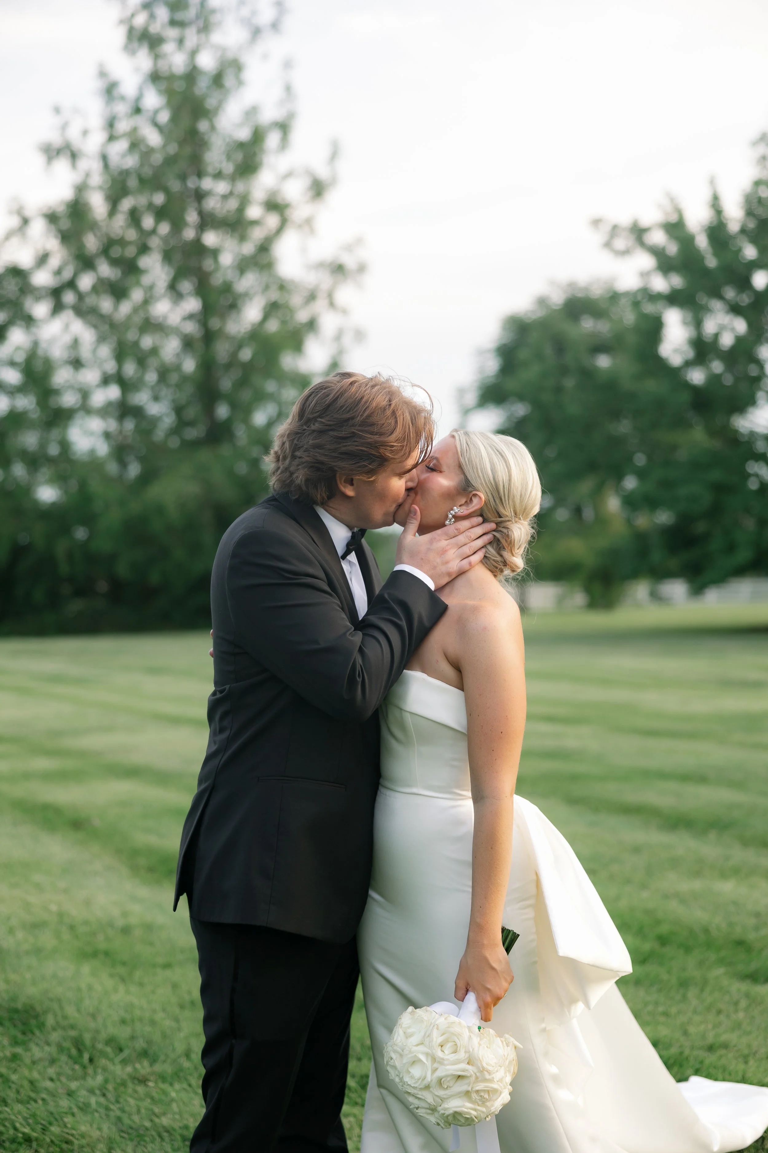 A newlywed couple kisses outdoors on a grassy field, with trees in the background. The groom is wearing a black tuxedo and the bride is in a strapless white wedding gown holding a bouquet of white roses.