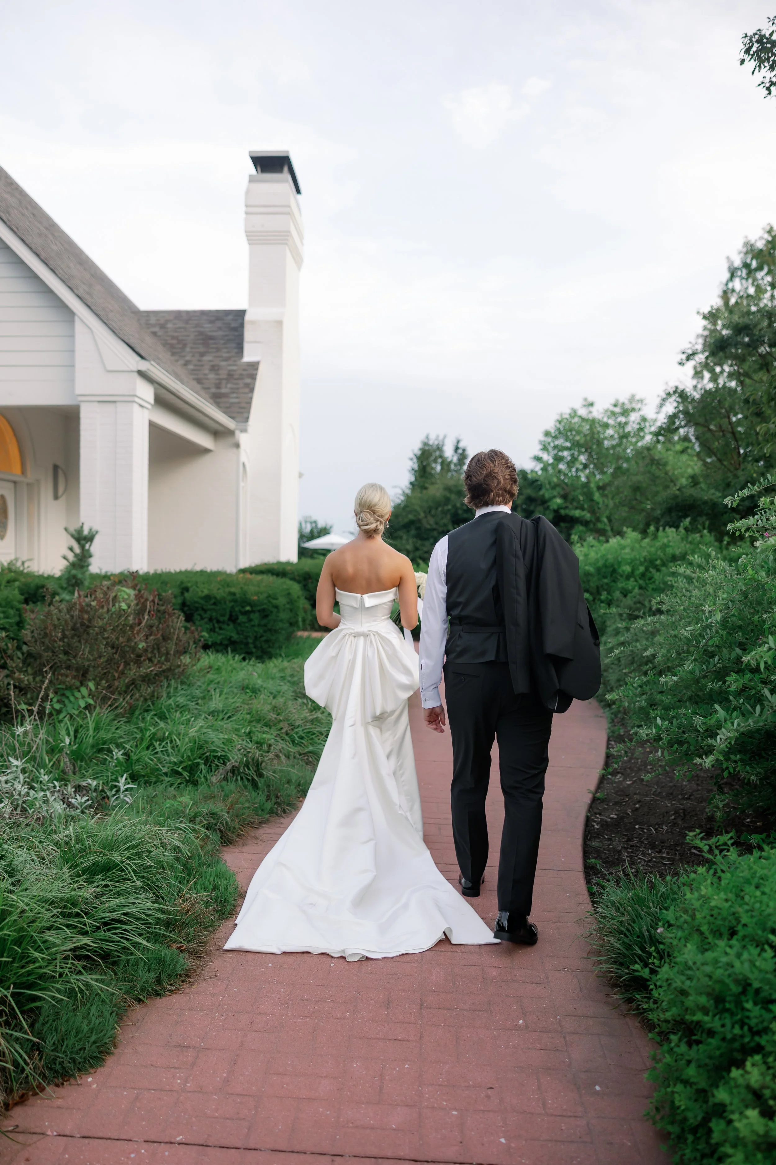 A bride and groom walk hand in hand along a brick pathway outside a white house with a chimney, surrounded by greenery.