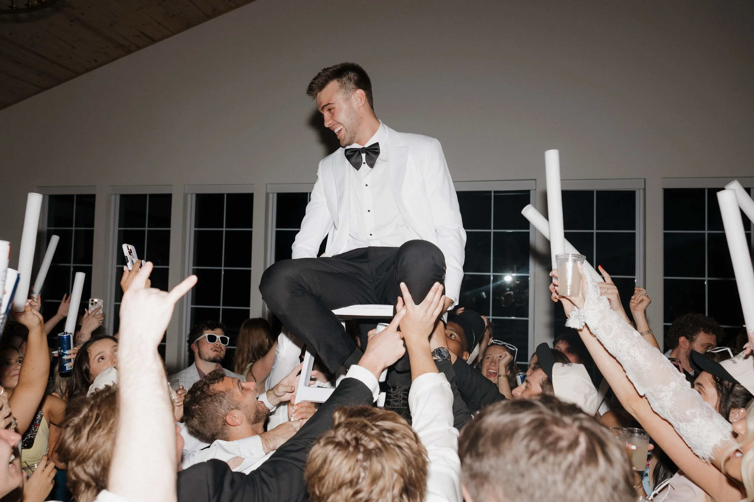A man dressed in a tuxedo and white jacket sitting on a chair held up by people at a party or celebration, surrounded by crowd celebrating and holding rolled papers and drinks.