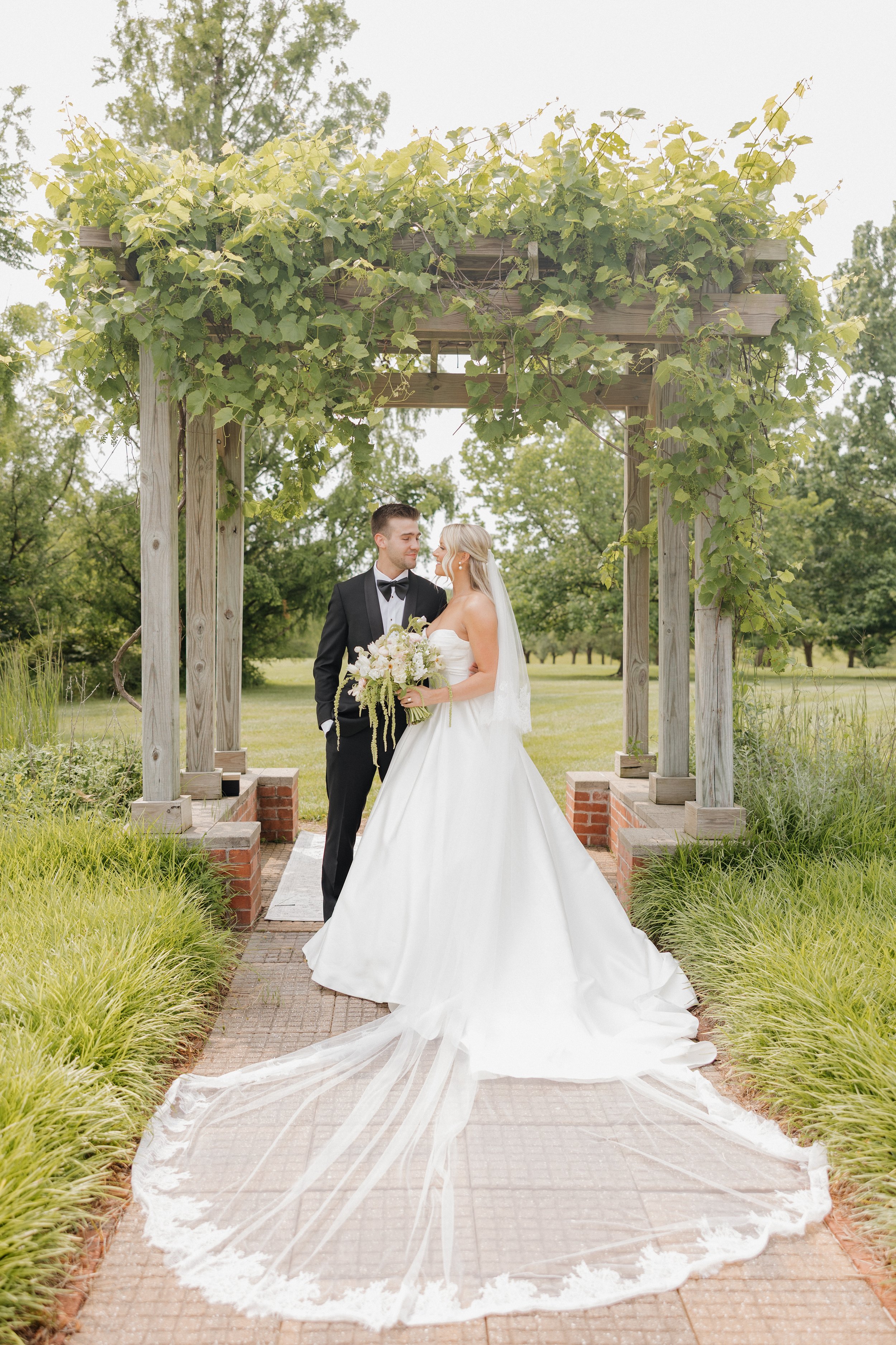 Bride and groom standing under a wooden archway covered with green vines, outdoors in a park-like setting, during their wedding.