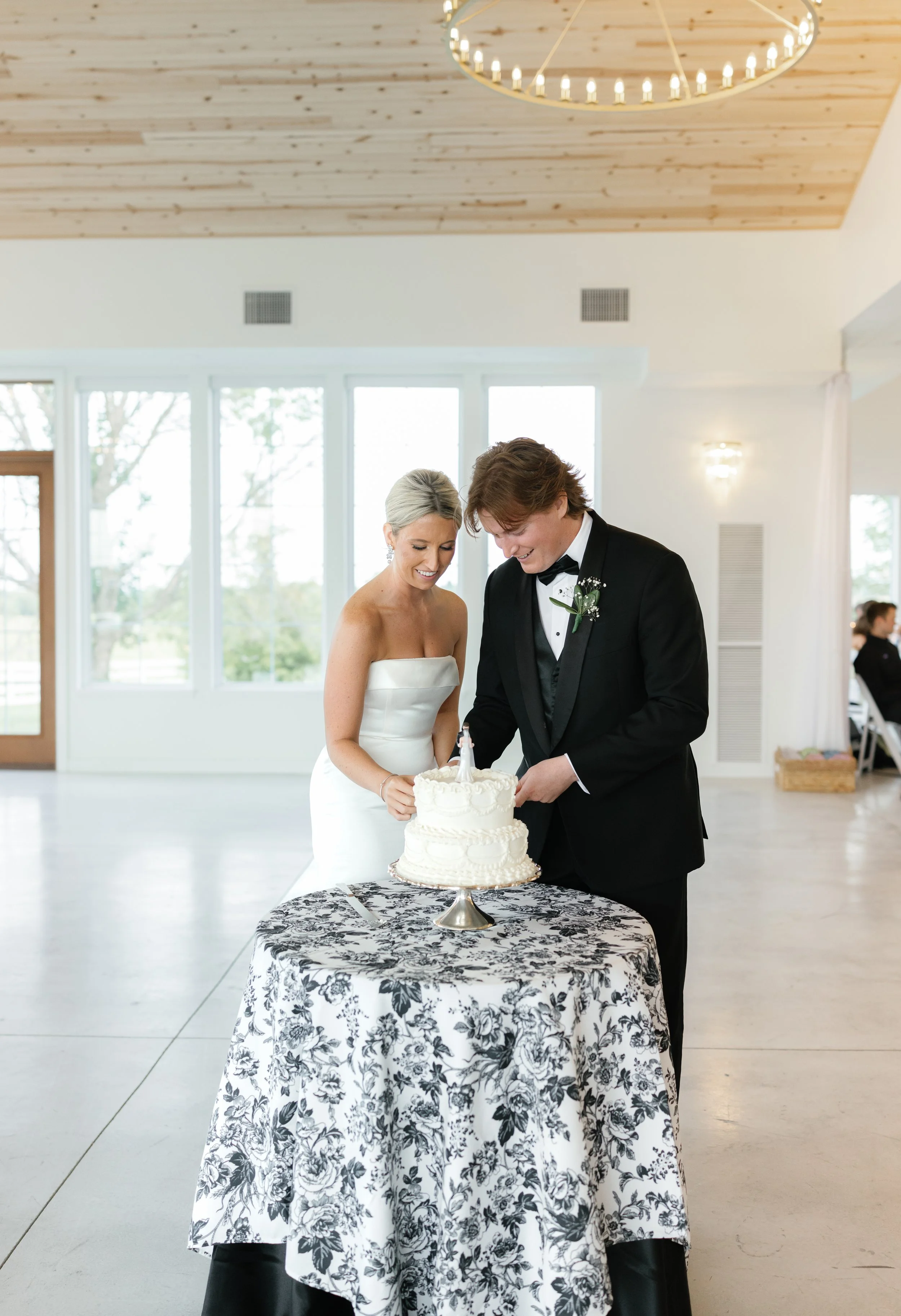 Bride and groom cutting wedding cake at reception indoors with large windows and wooden ceiling.
