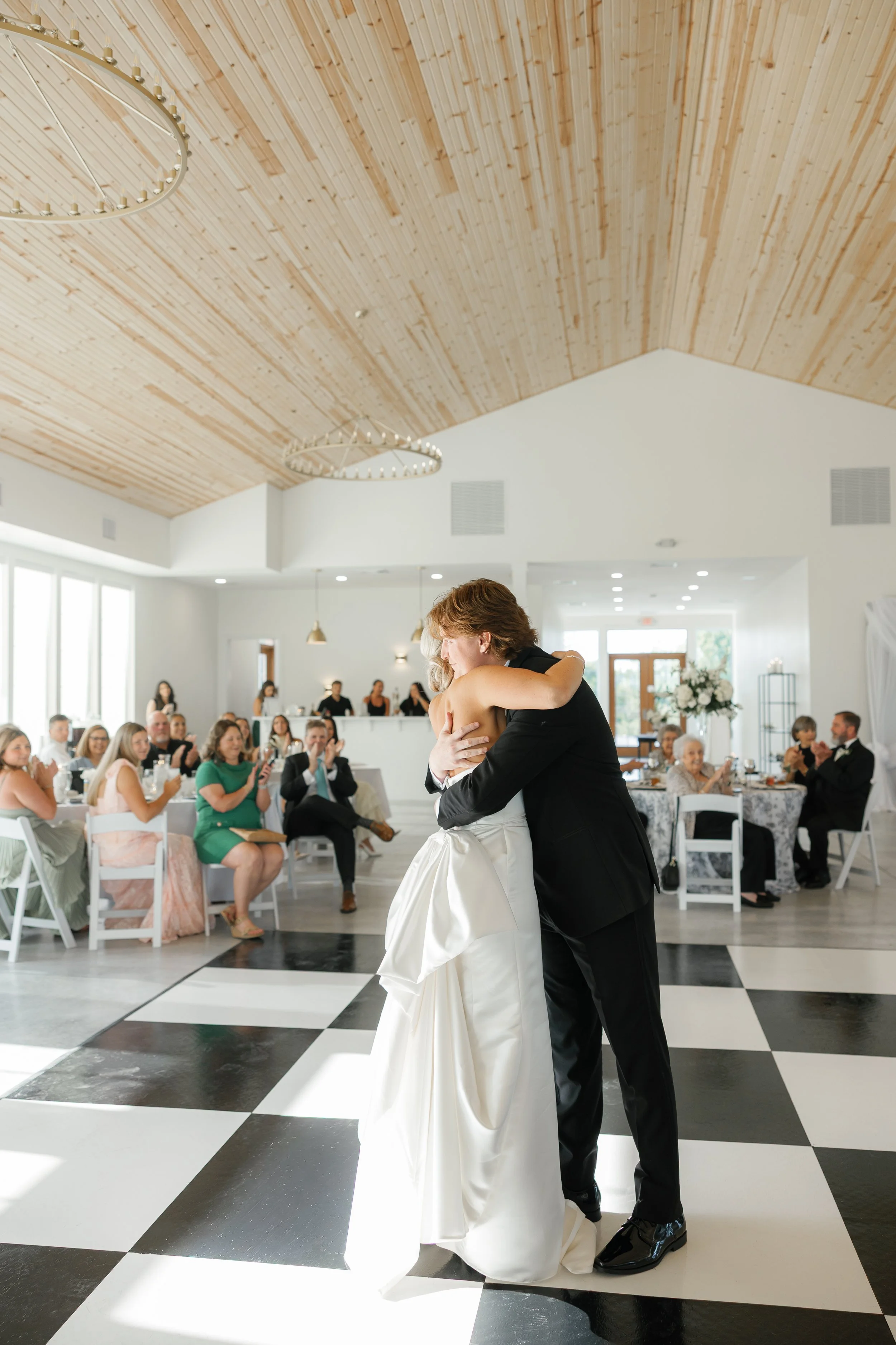 A bride and groom share a dance during their wedding reception, with guests seated at tables watching and clapping in a bright, modern venue with a wooden ceiling and black-and-white checkered dance floor.