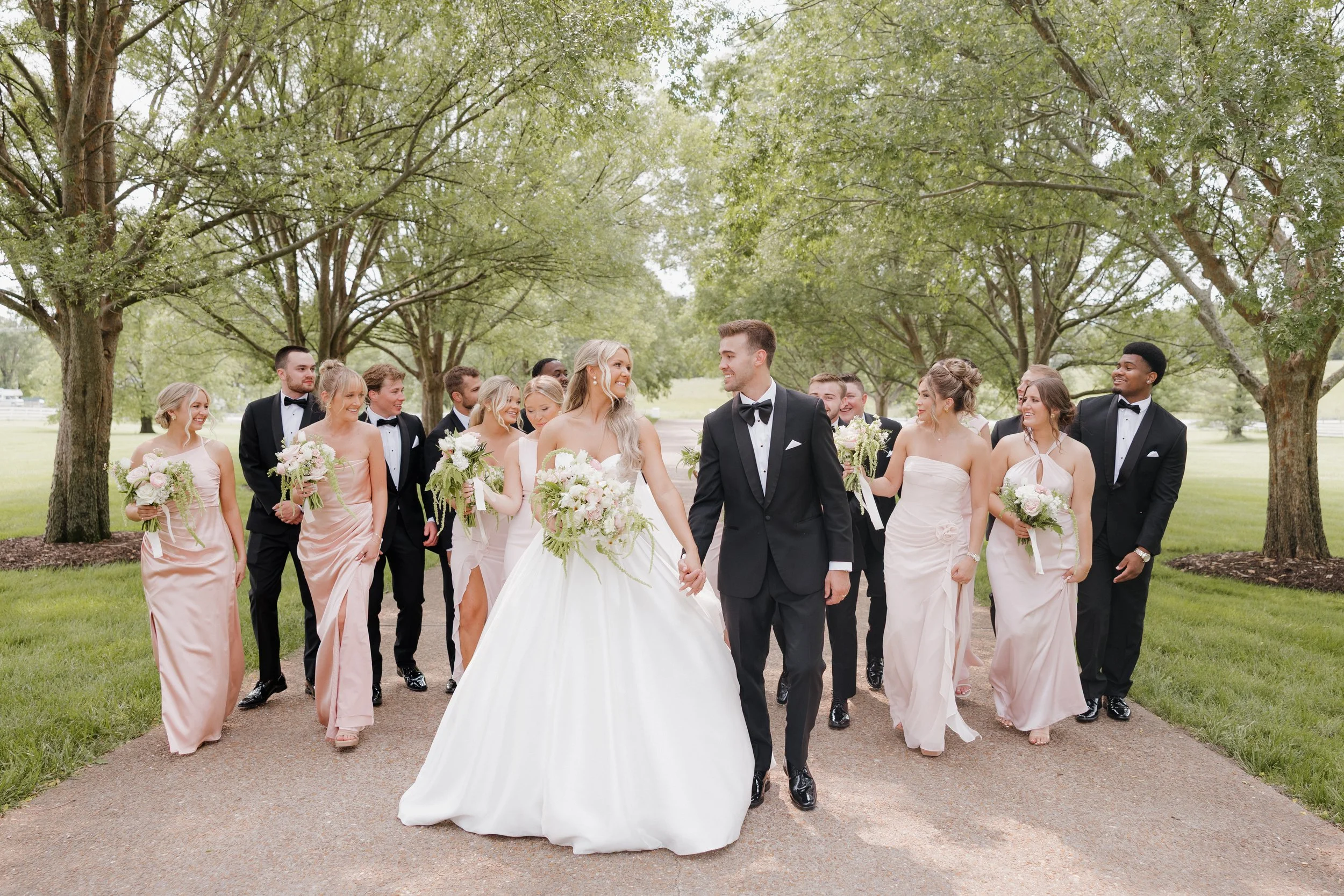 A bride and groom walking hand in hand with their wedding party outdoors on a tree-lined pathway during the daytime.