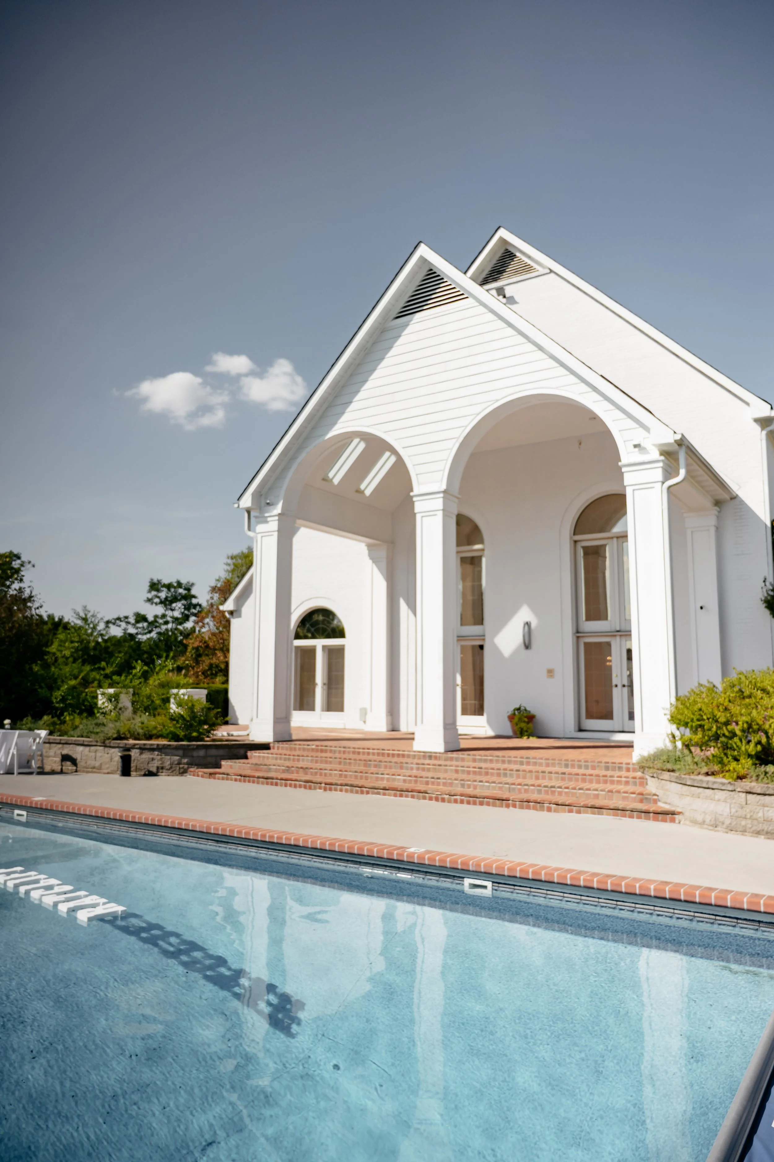 White house with arched porch, brick steps, and swimming pool in the foreground.