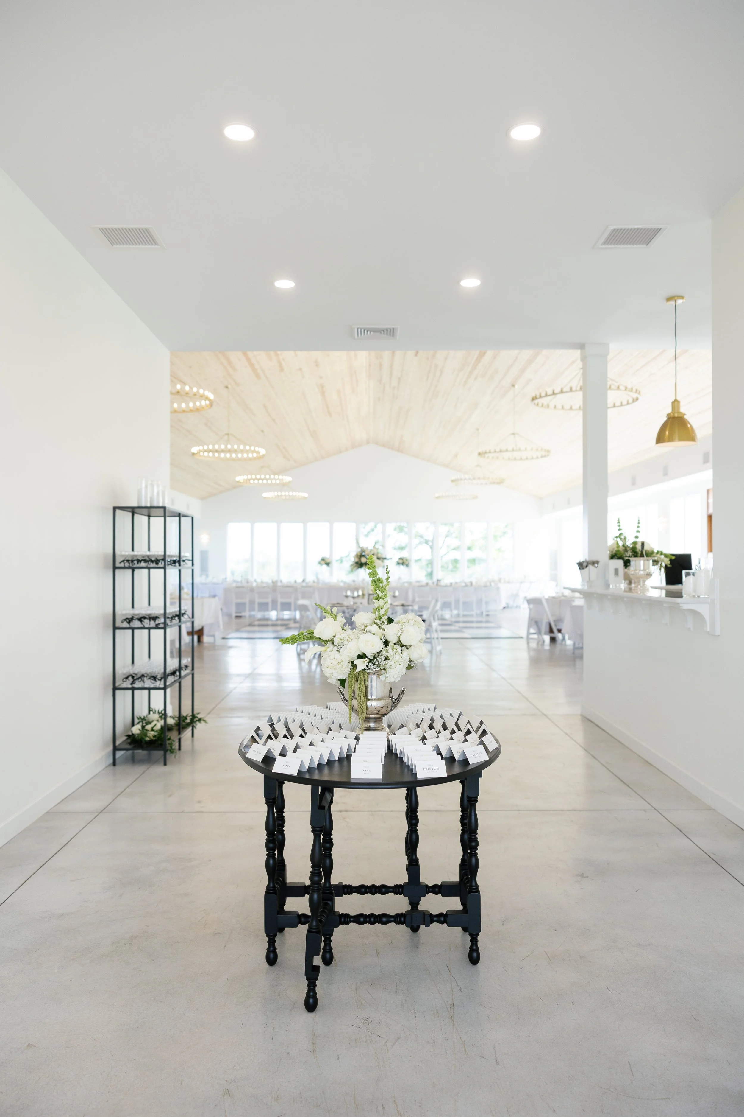 A reception or event space with a black table in the foreground, decorated with white flowers in a silver vase and place cards. The space features large windows, circular chandeliers, and a minimalist design with white walls and a wooden ceiling.