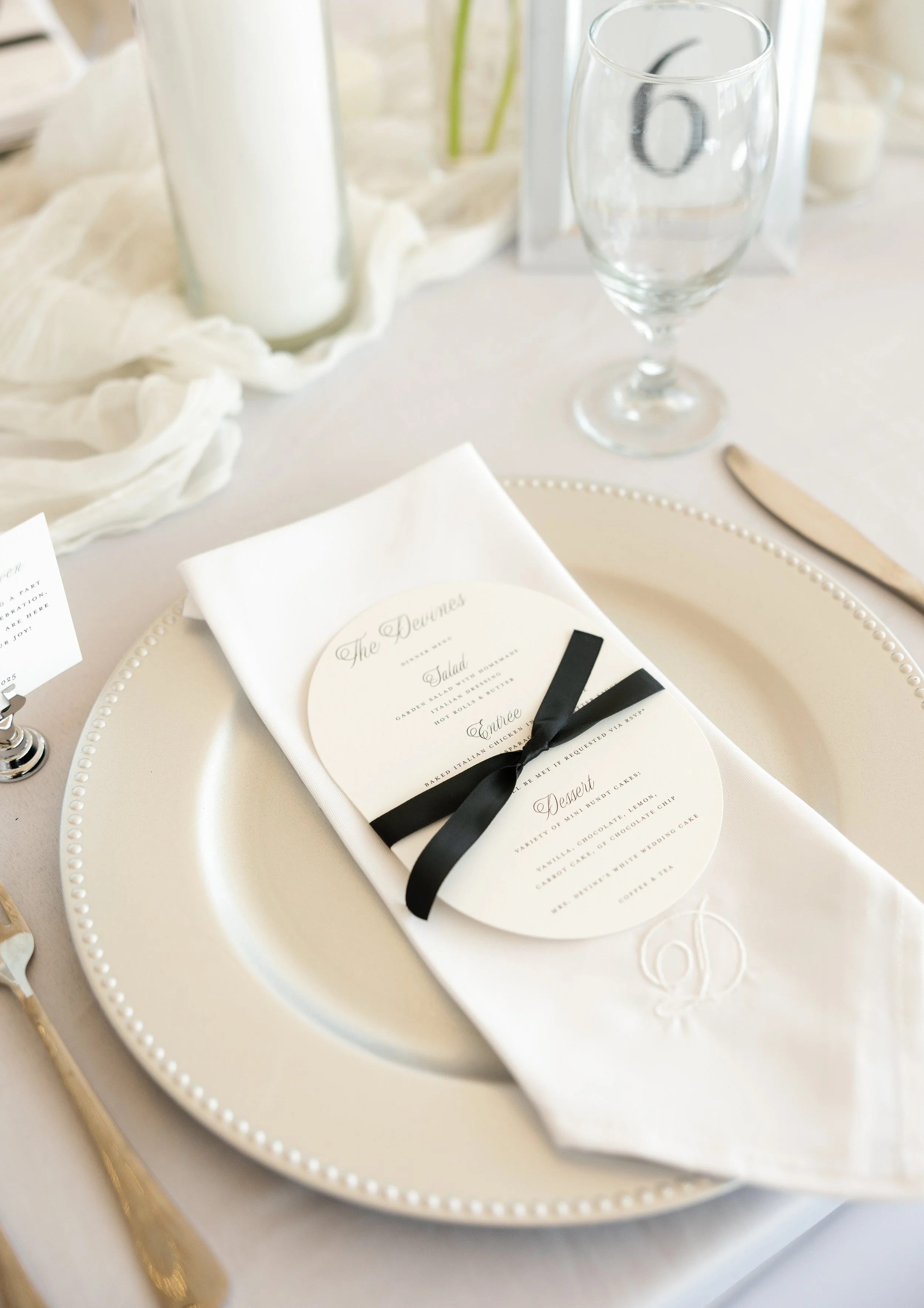Elegant table setting with a white plate, a white napkin with an initial embroidered in white, a menu tied with a black ribbon, a clear wine glass, and gold flatware at a formal event.