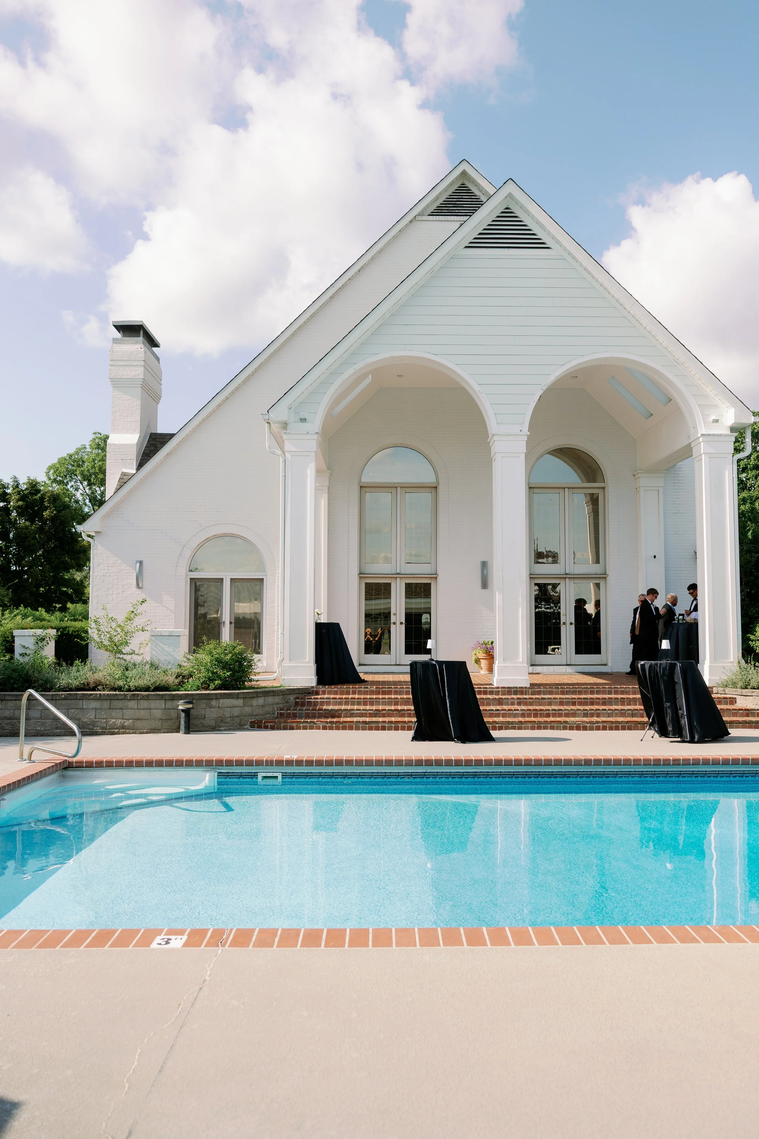A white house with a pitched roof, arched windows and doors, and a chimney, situated behind a swimming pool with a blue water surface, under a partly cloudy sky. There are small groups of people dressed in formal attire gathered near the house entran