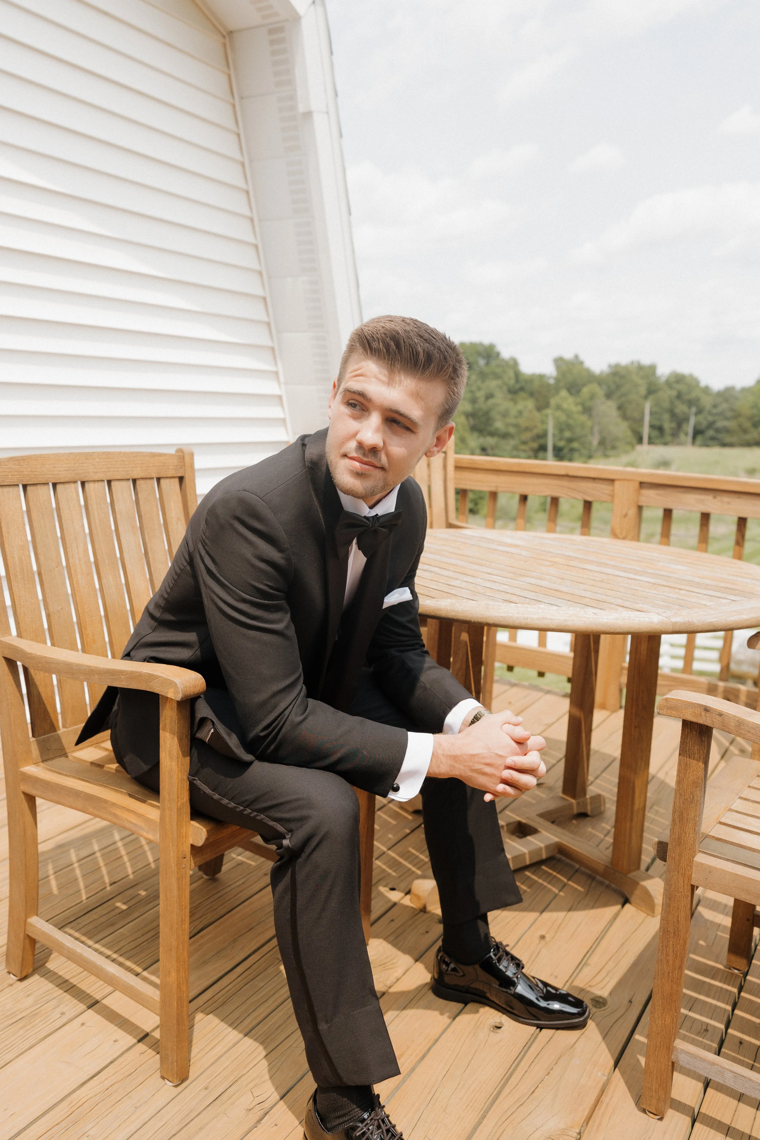 A young man in a black tuxedo with a bow tie, sitting on a wooden outdoor deck with matching wooden furniture, looking thoughtful with a serious expression.