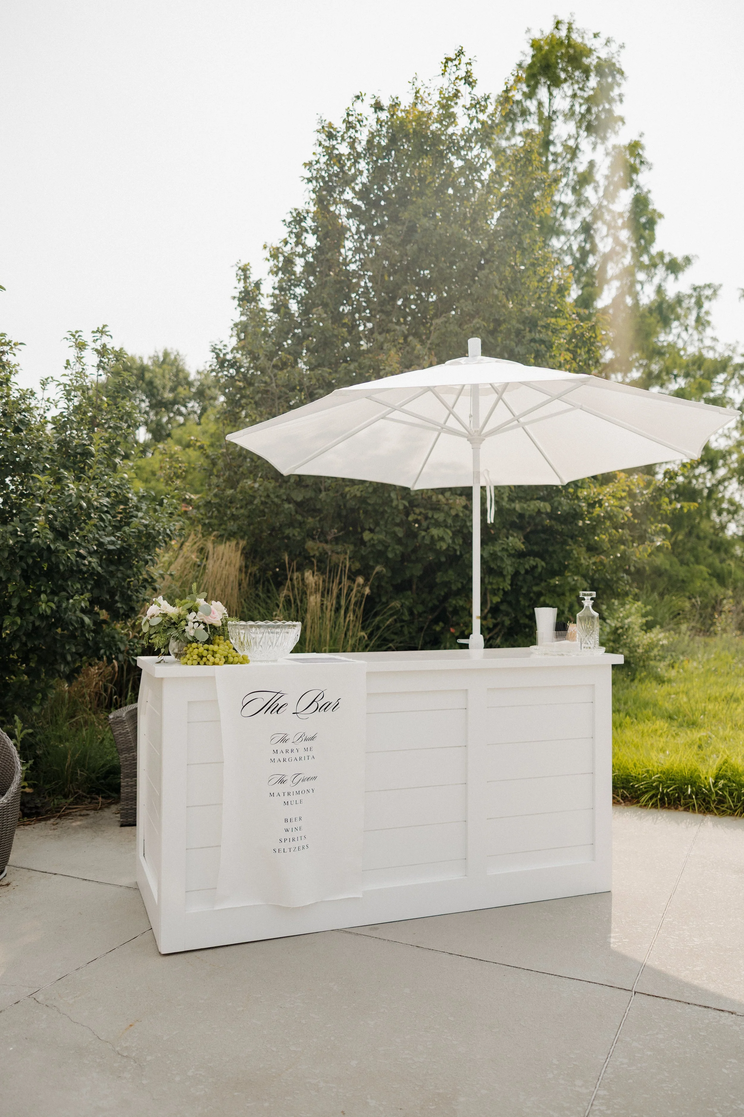 A white outdoor bar with a large white umbrella, floral arrangement, glassware, and bottles, set against a background of green trees and grass.