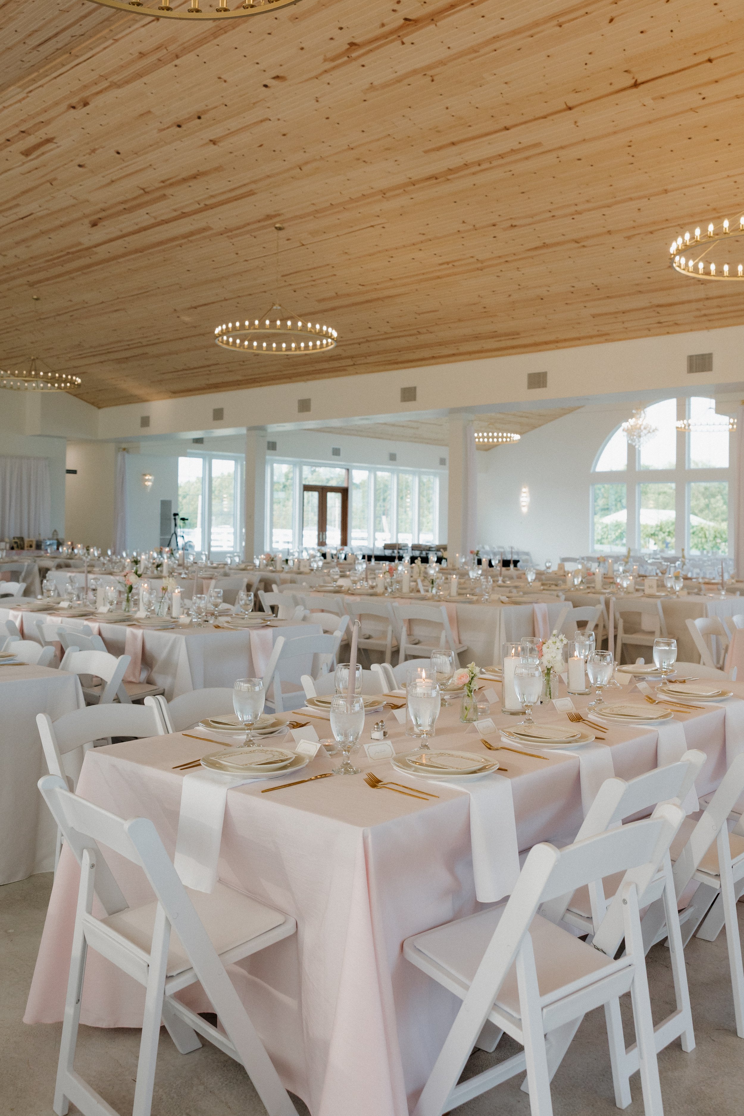 Wedding reception hall with white tables and chairs, decorated with floral centerpieces, candles, and gold utensils, featuring a wooden ceiling and large windows letting in natural light.