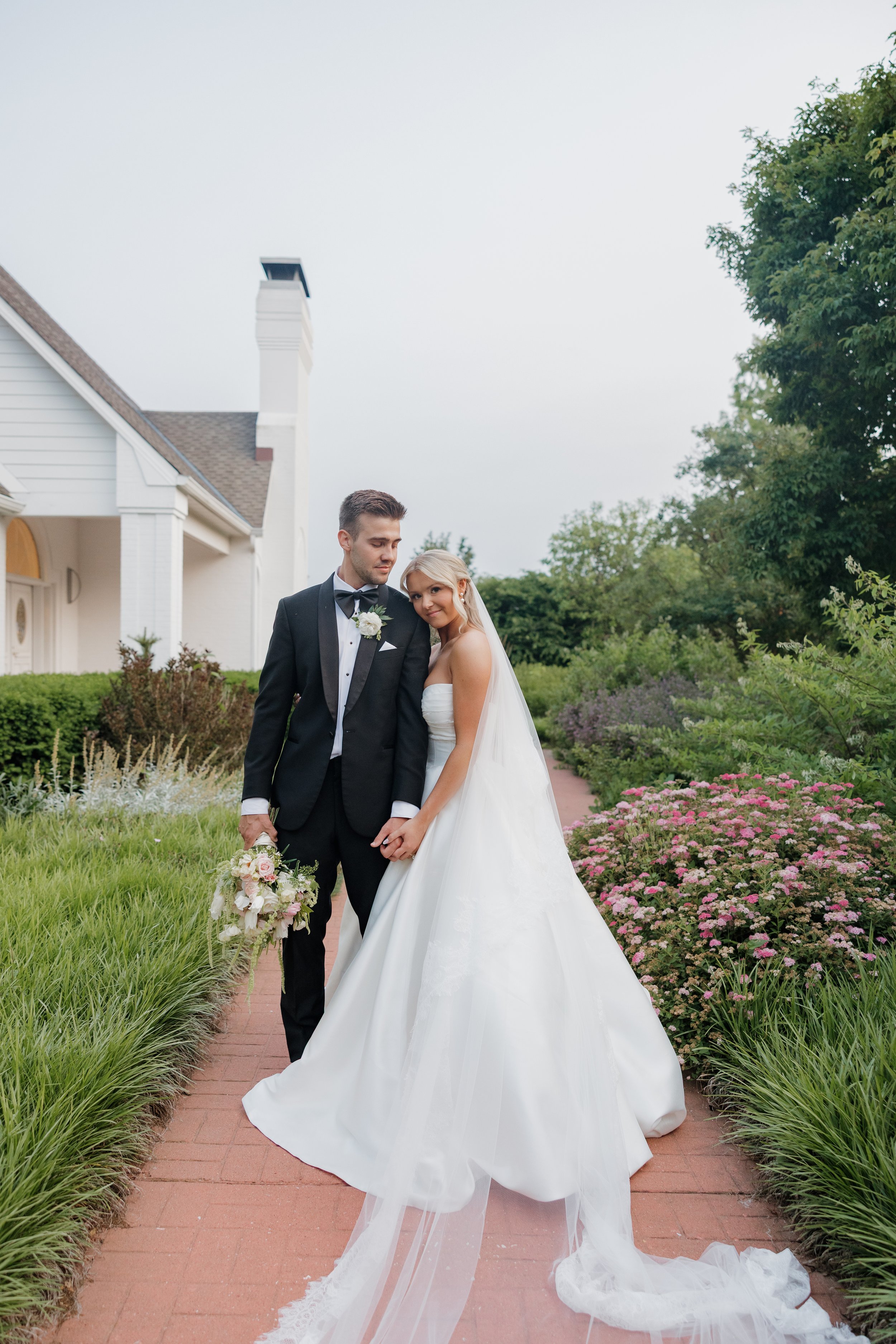 A bride and groom standing on a brick pathway outdoors, with the bride in a white wedding gown and veil, and the groom in a black tuxedo holding a bouquet, surrounded by greenery and pink flowers.