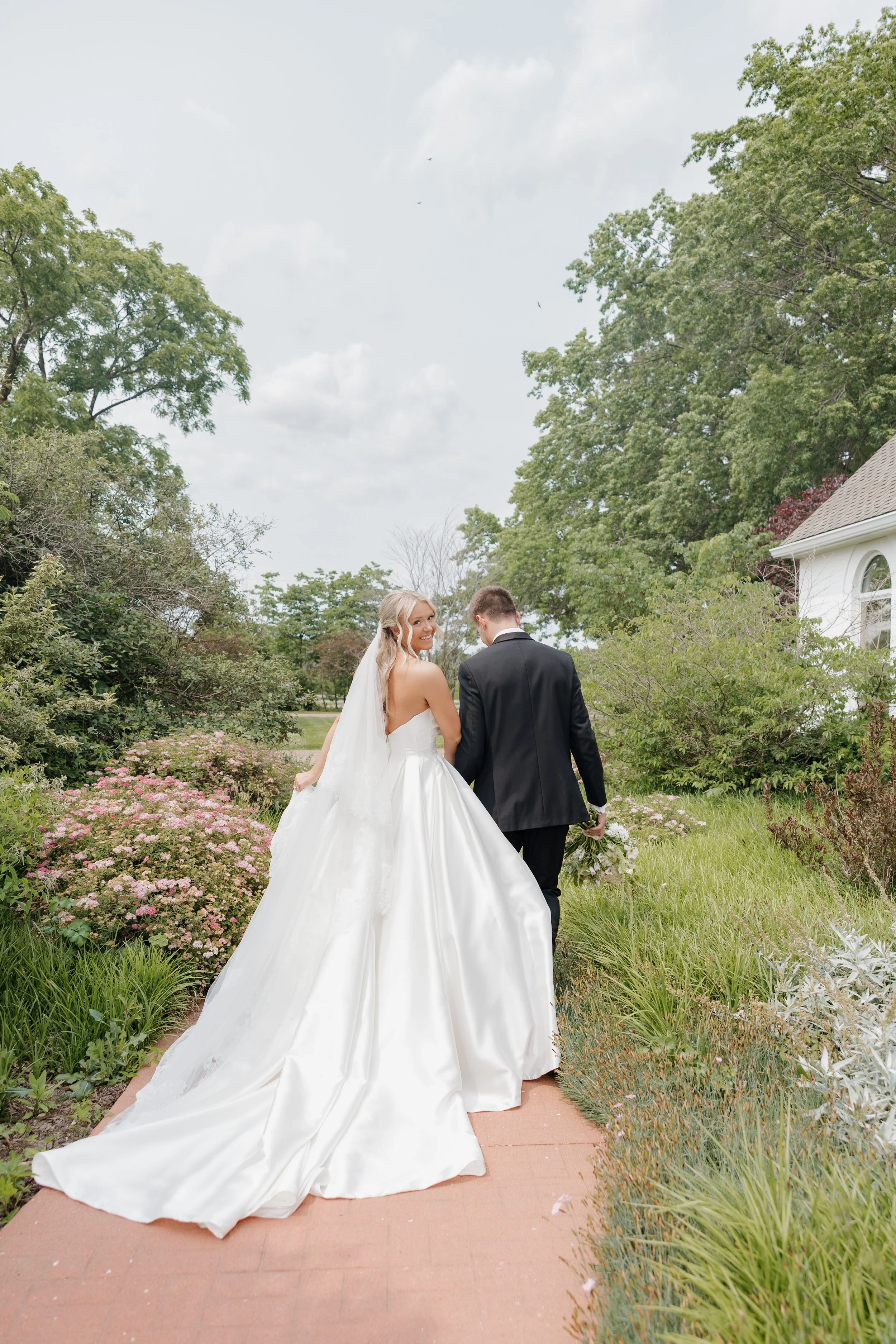 Bride and groom walking on a garden path, with the bride in a white wedding gown and the groom in a black suit, surrounded by greenery and flowers.
