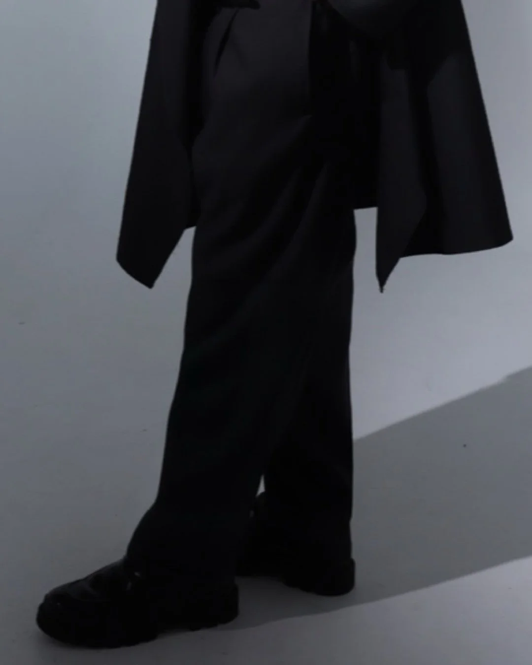 Person dressed in black clothing standing against a minimalist gray background.