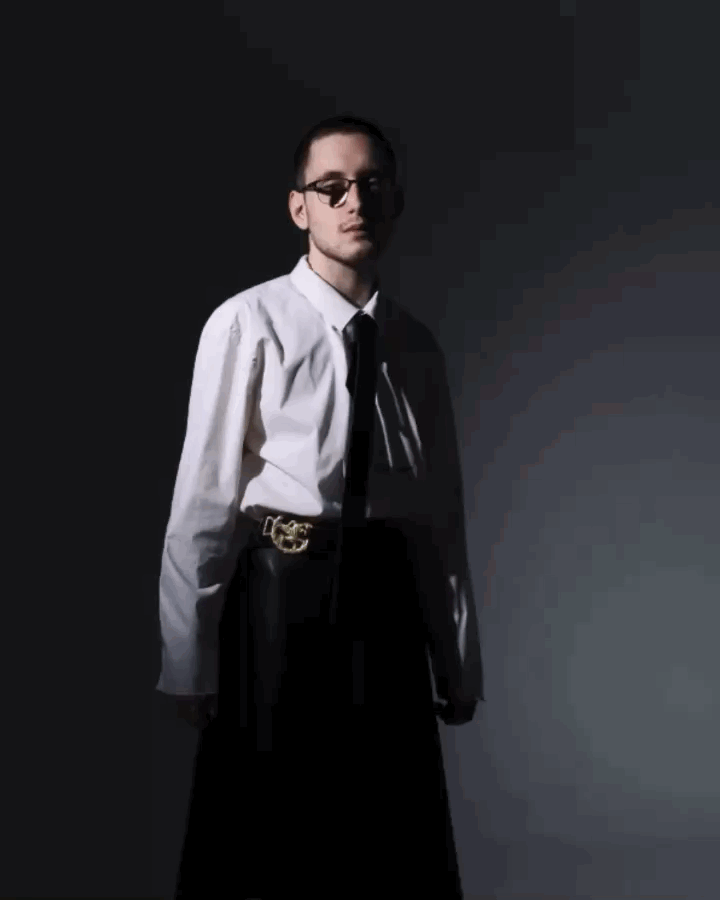 A young man with glasses wearing a white shirt, black tie, and black skirt with a decorative belt, standing against a dark gradient background.