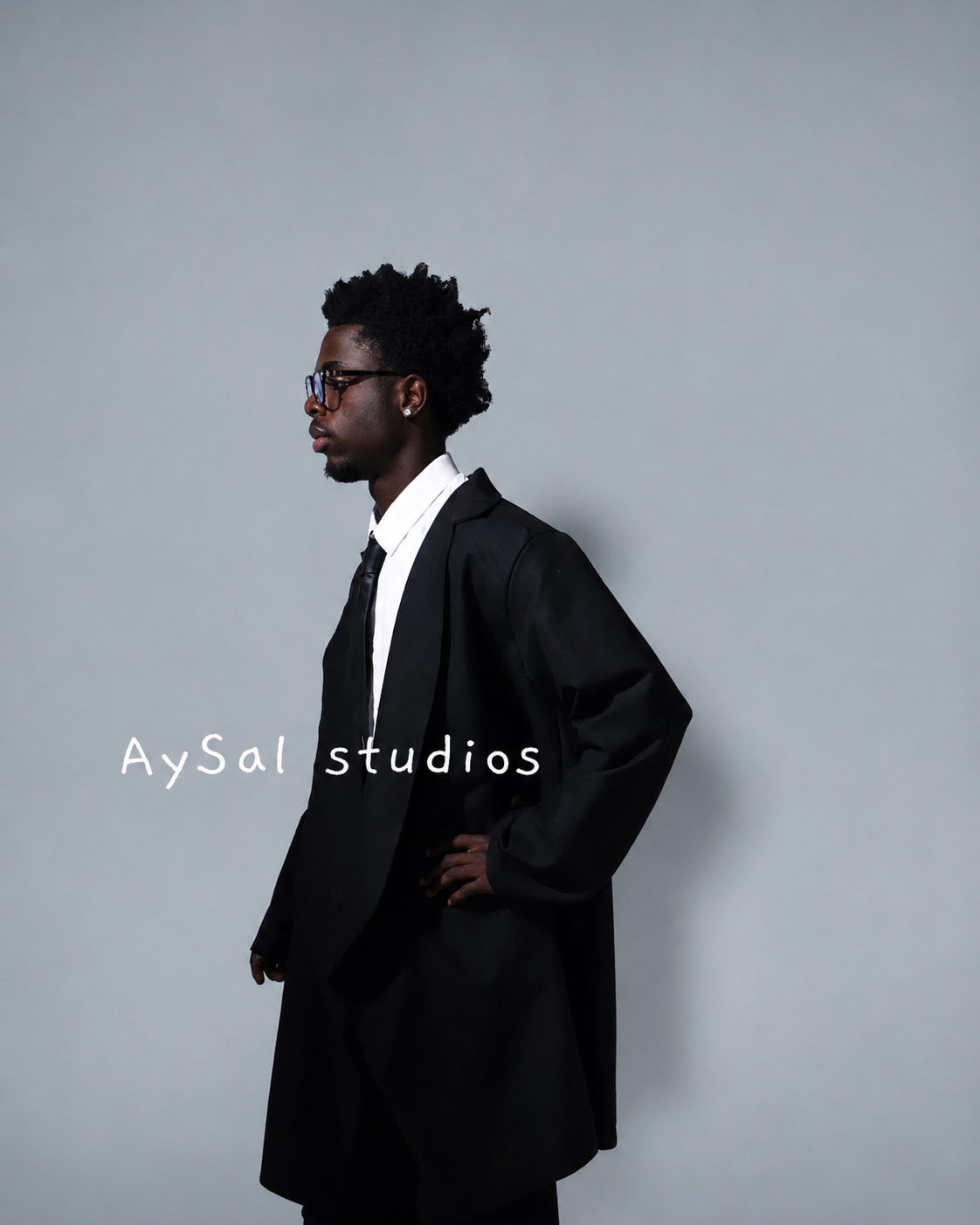 A man dressed in a black suit with a white shirt and black tie, wearing glasses, standing in profile against a plain gray background, with the text 'AySal studios' on the lower left.