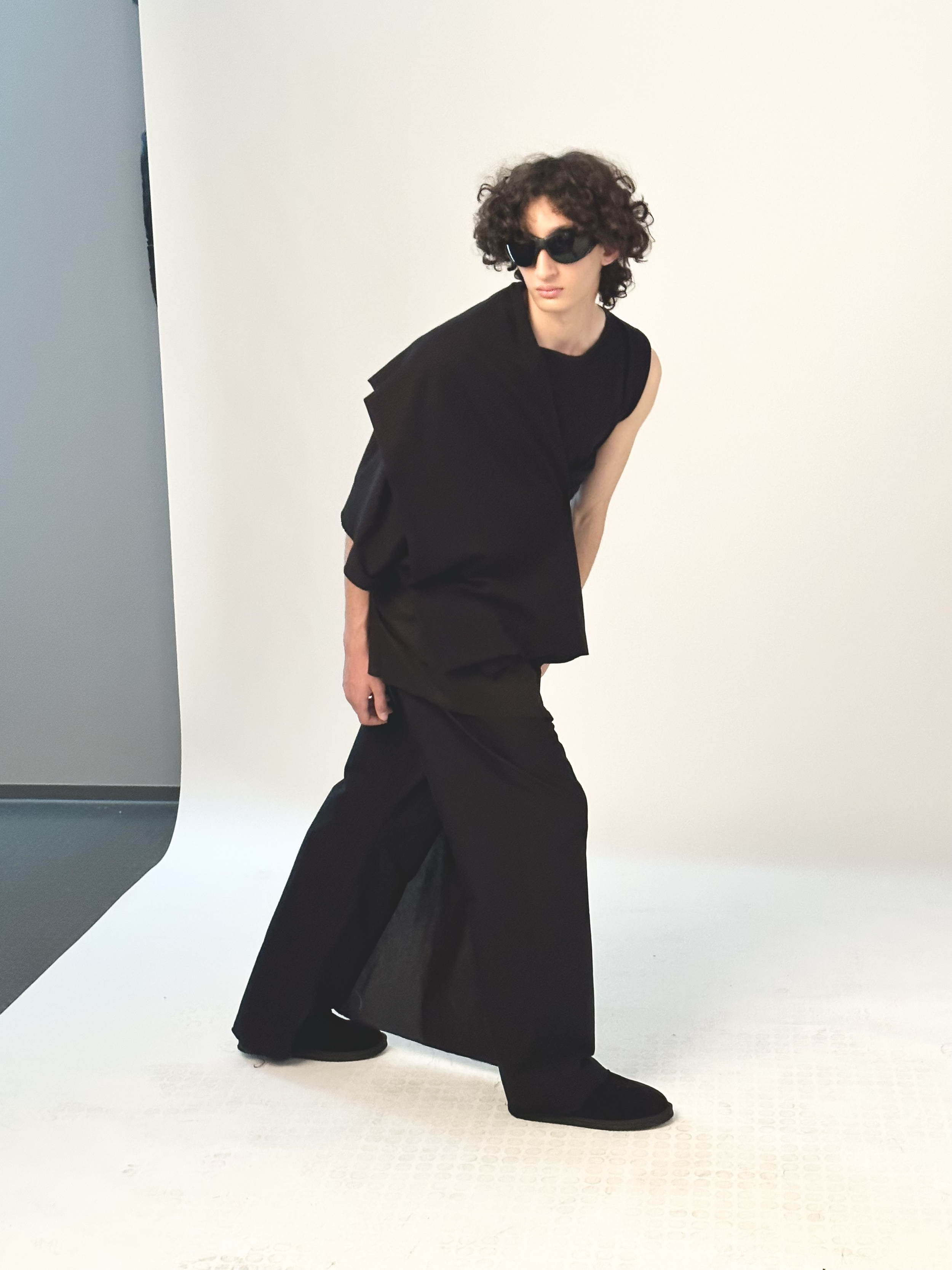 Person with curly hair wearing black sunglasses, a sleeveless black top, wide-leg black pants, and black shoes, posing against a plain backdrop.