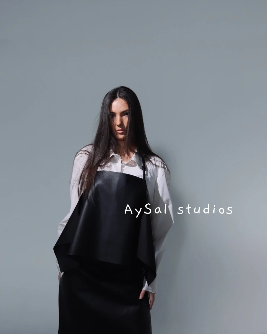 A woman with long dark hair wearing a white shirt with a black apron, standing against a plain gray background with the text 'AySal studios'.