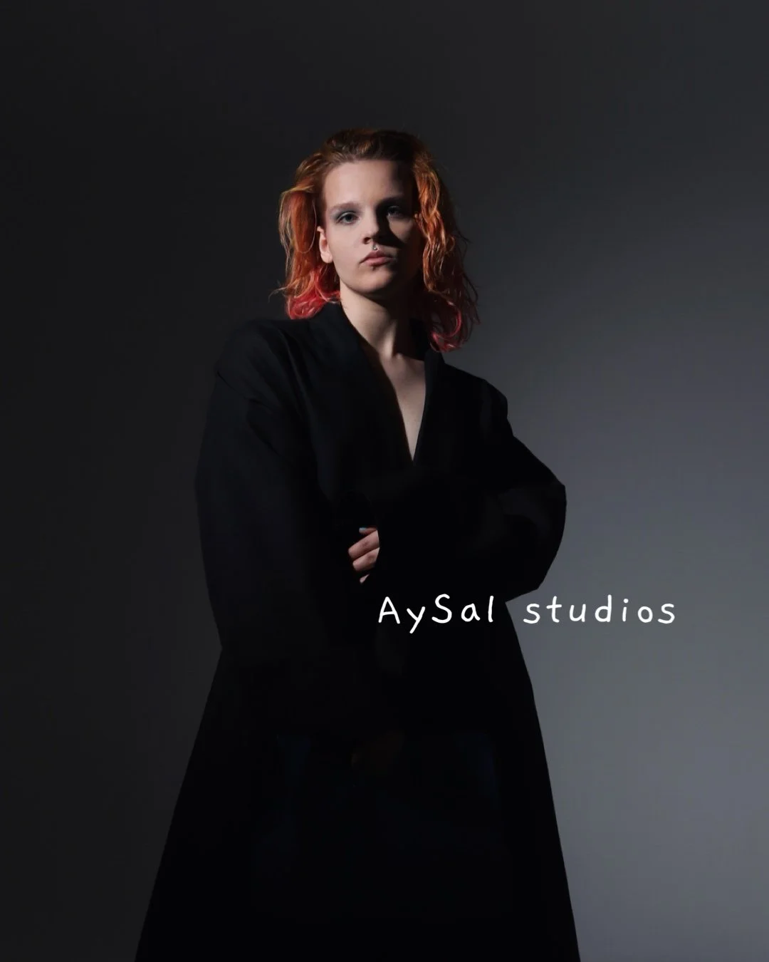 A woman with red hair, wearing a black jacket, posing with arms crossed against a dark gradient background. Text overlay reads 'AySal studios'.