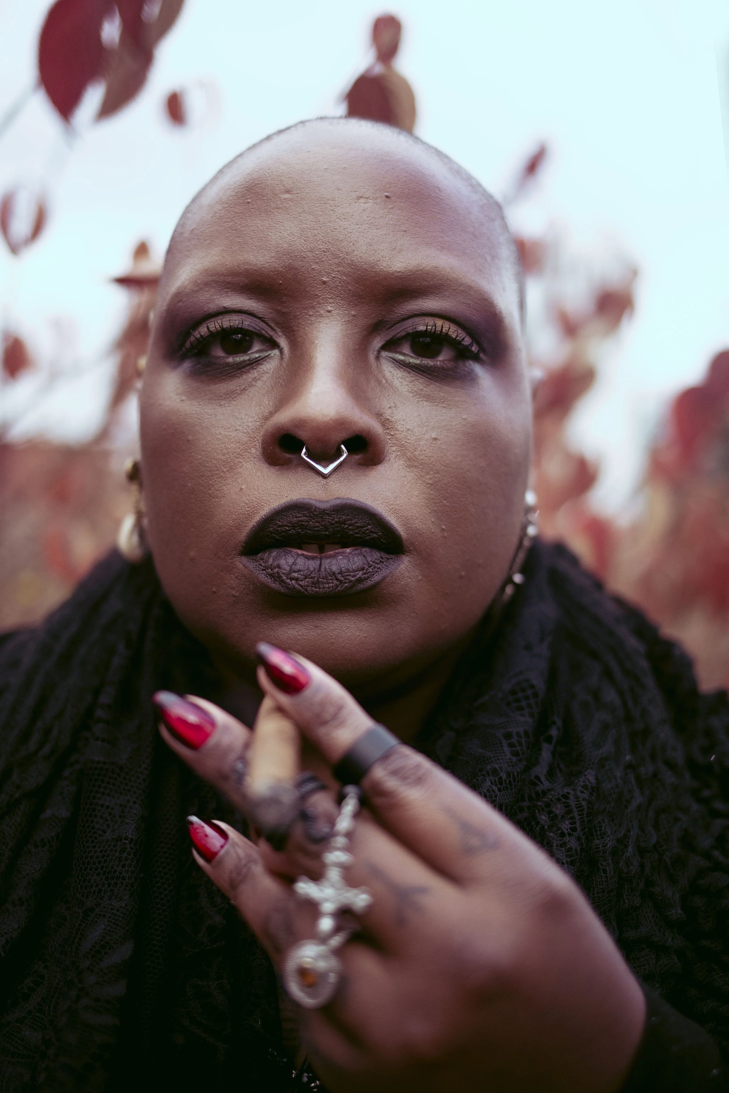 A close-up portrait of Samar Jade with a shaved head, dark makeup, jewelry, and rings, with red and brown leaves in the background.