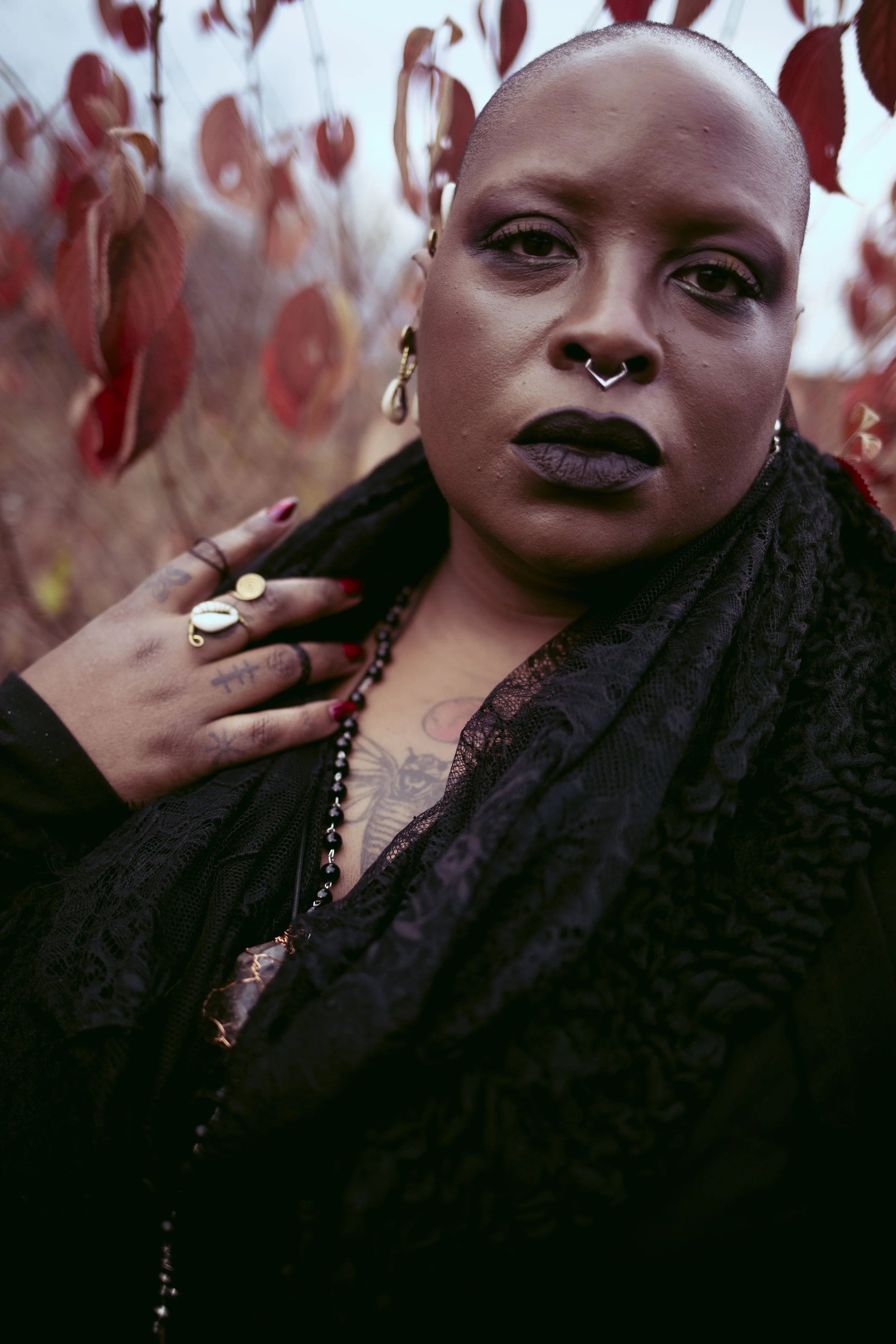 Samar Jade with dark lipstick and piercings, wearing black clothing and jewelry, posed outdoors with red leaves in the background.