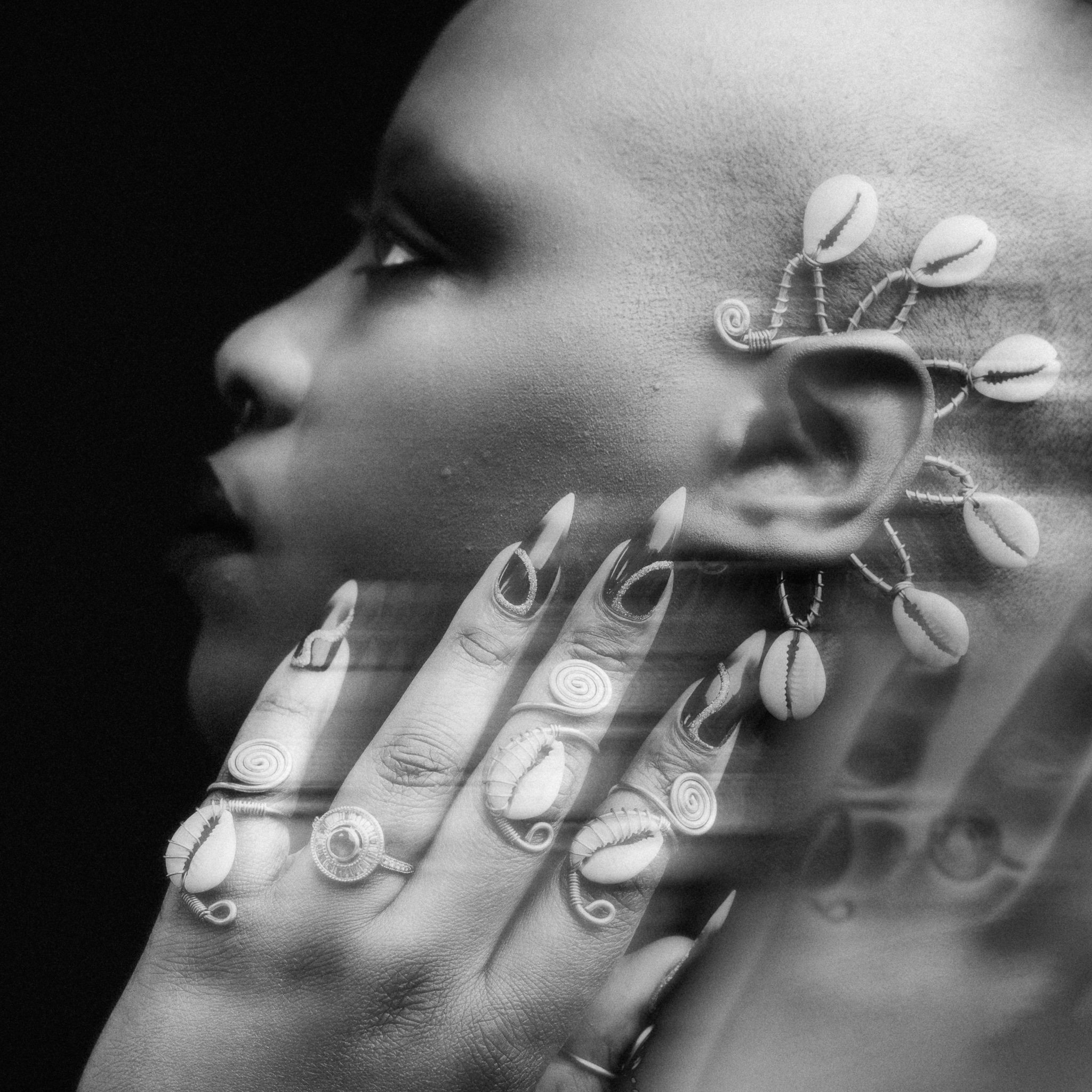 A person with short hair and makeup showcases jewelry on their face, hand, and fingers, including rings and a necklace with shell and wire designs.