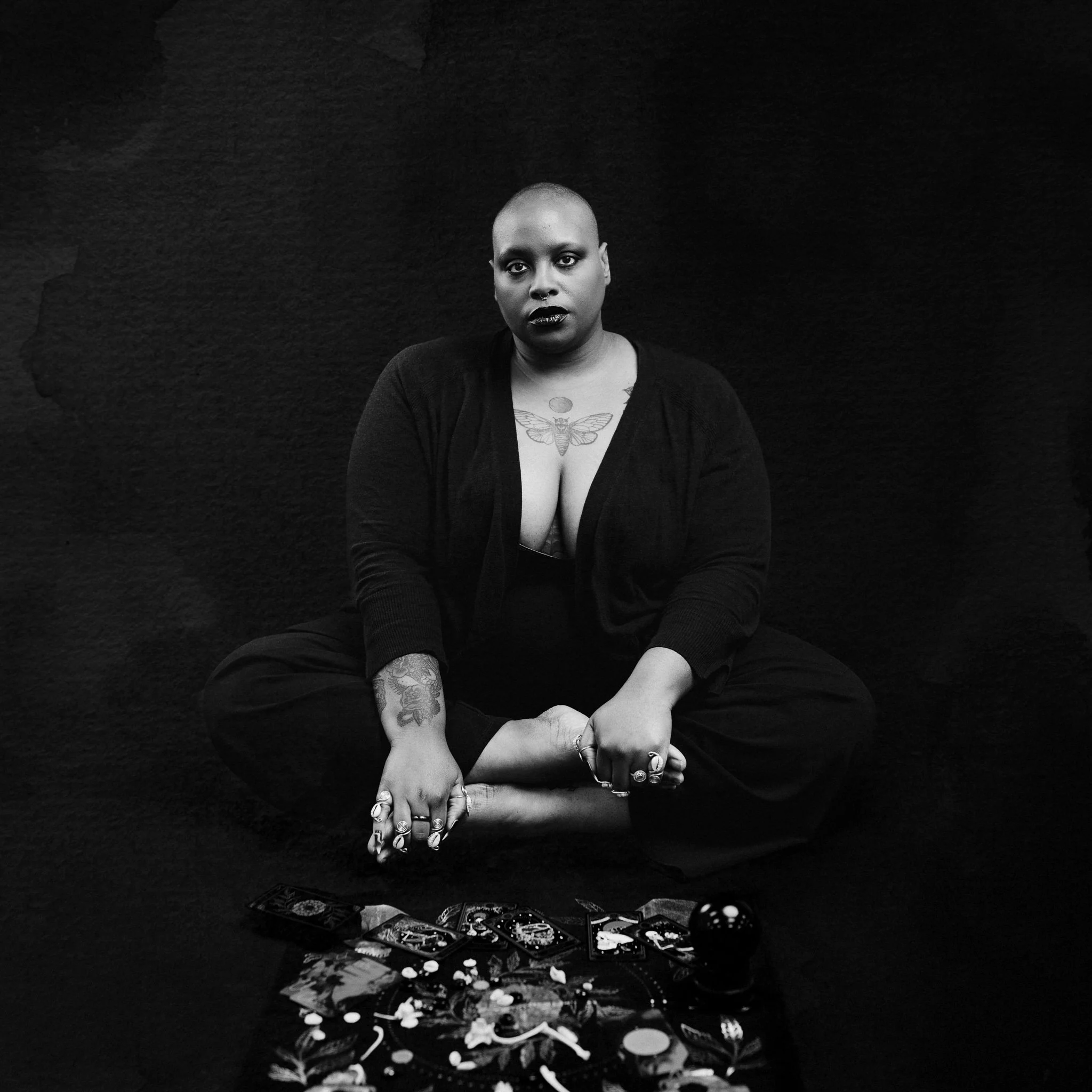 A tattooed human with a shaved head sitting cross-legged on the floor with tarot cards and various objects in front of them, against a dark background.