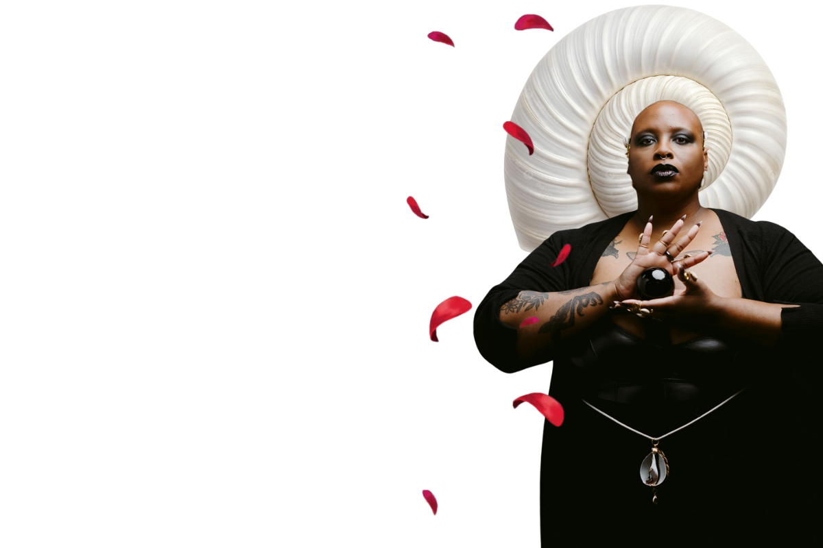 A human with dark makeup and tattoos, holding a black sphere, is positioned in front of a large, white, spiral-shaped headpiece with pink accents, against a black background.