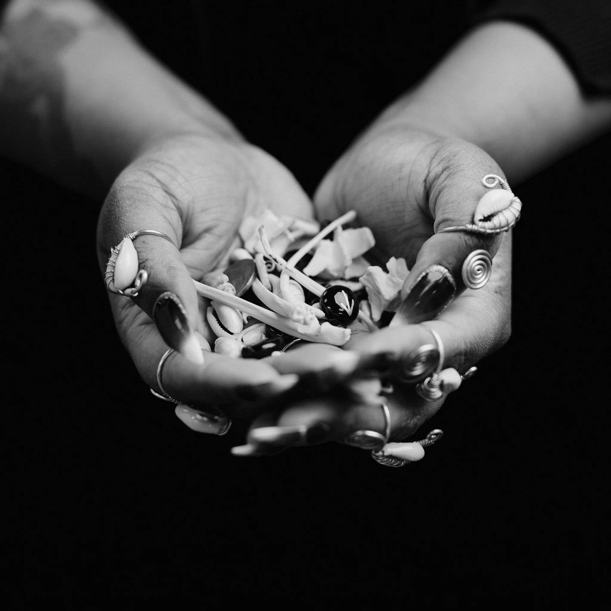 A pair of hands with multiple rings holding a handful of seashells, small objects, and candies in black and white.