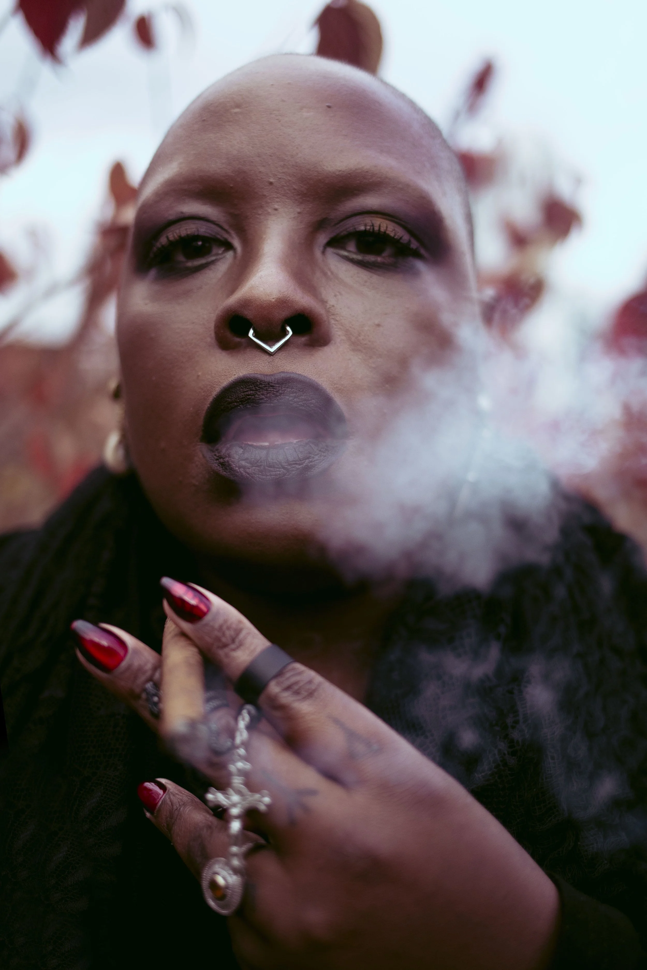 Samar Jade with dark skin, shaved head, and dark lipstick smoking, with smoke visible near their face, wearing rings and jewelry, and with a serious expression.