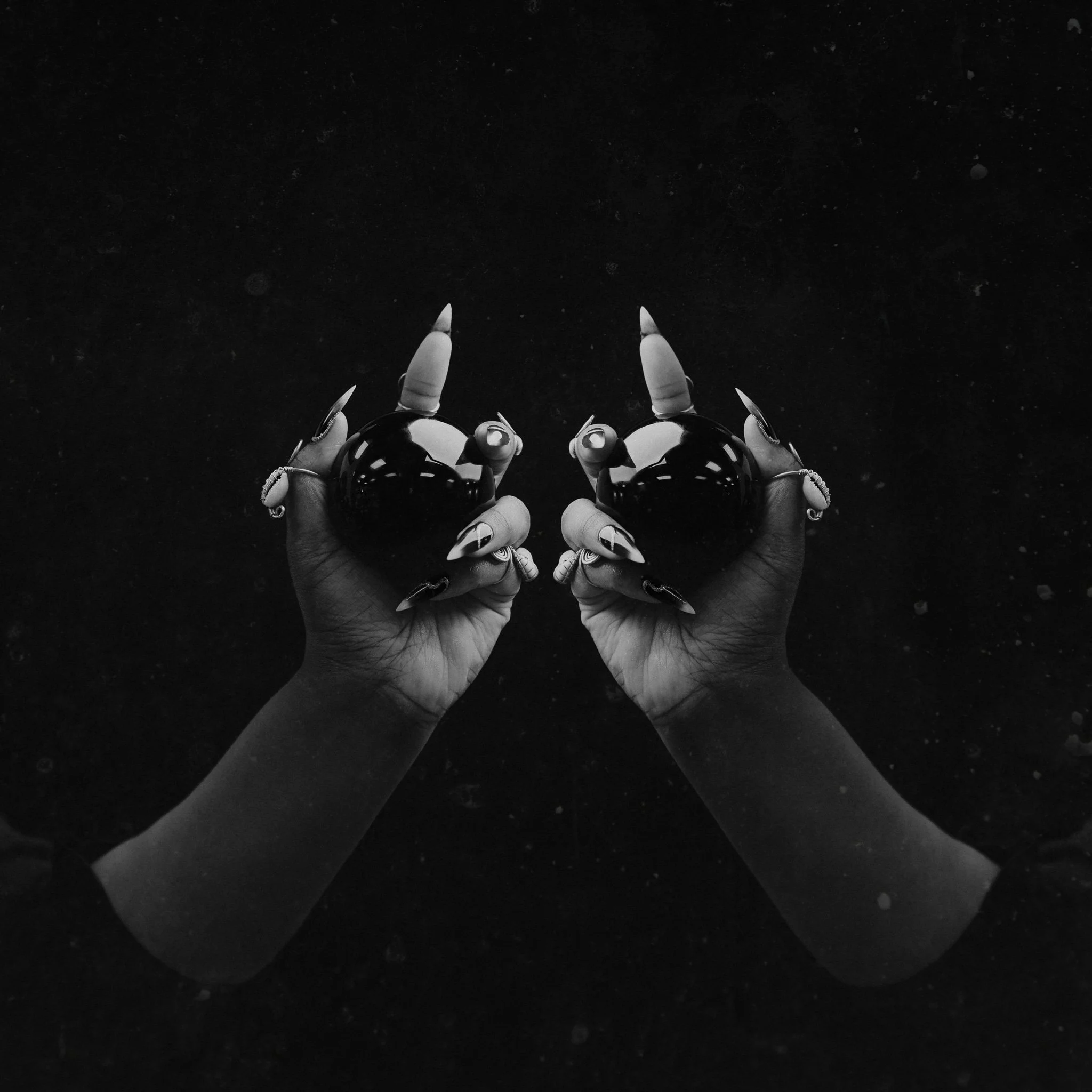 Black and white photograph of two hands with long, pointed nails holding two reflective spheres, mirror images of each other, against a dark, textured background.