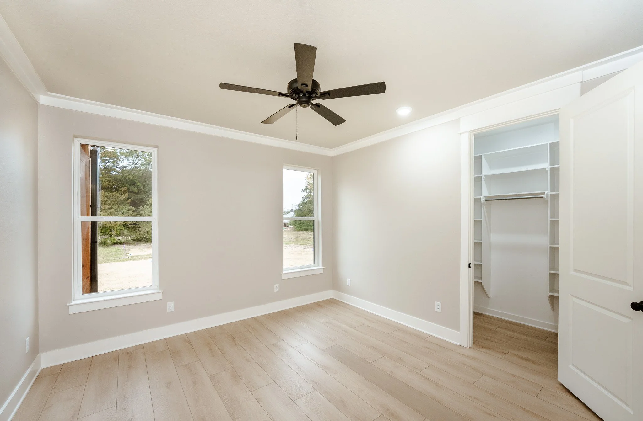 Empty bedroom with two windows, a ceiling fan, light wood flooring, and an open closet with white shelving.