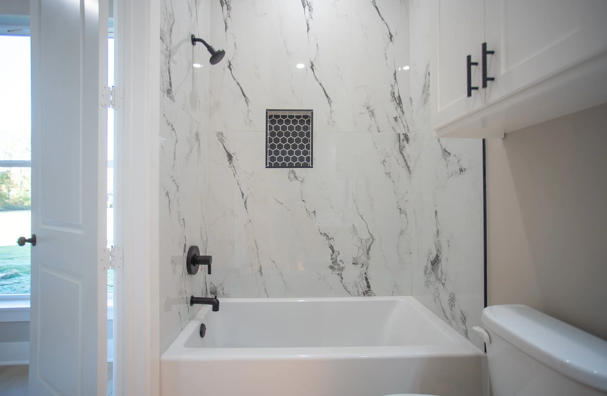Modern bathroom with white marble walls, a black showerhead, black faucet, and a white bathtub. There is a window with blinds and a cabinet on the right side.