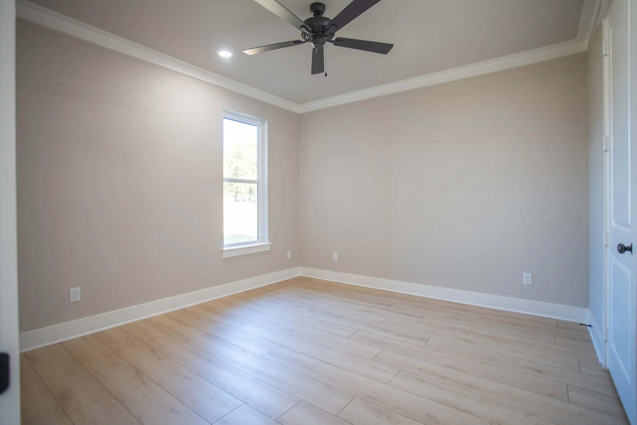 Empty room with beige walls, light wood floor, white trim, window, ceiling fan, and door.