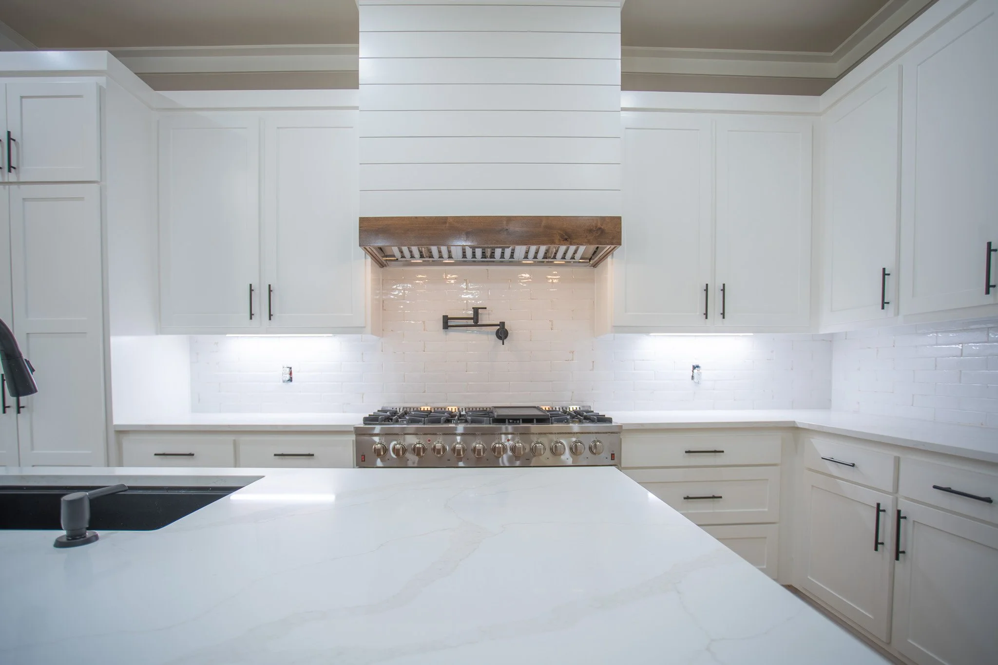 Modern kitchen with white cabinets, white marble countertops, a stainless steel stove, and a white brick backsplash under a wooden range hood.