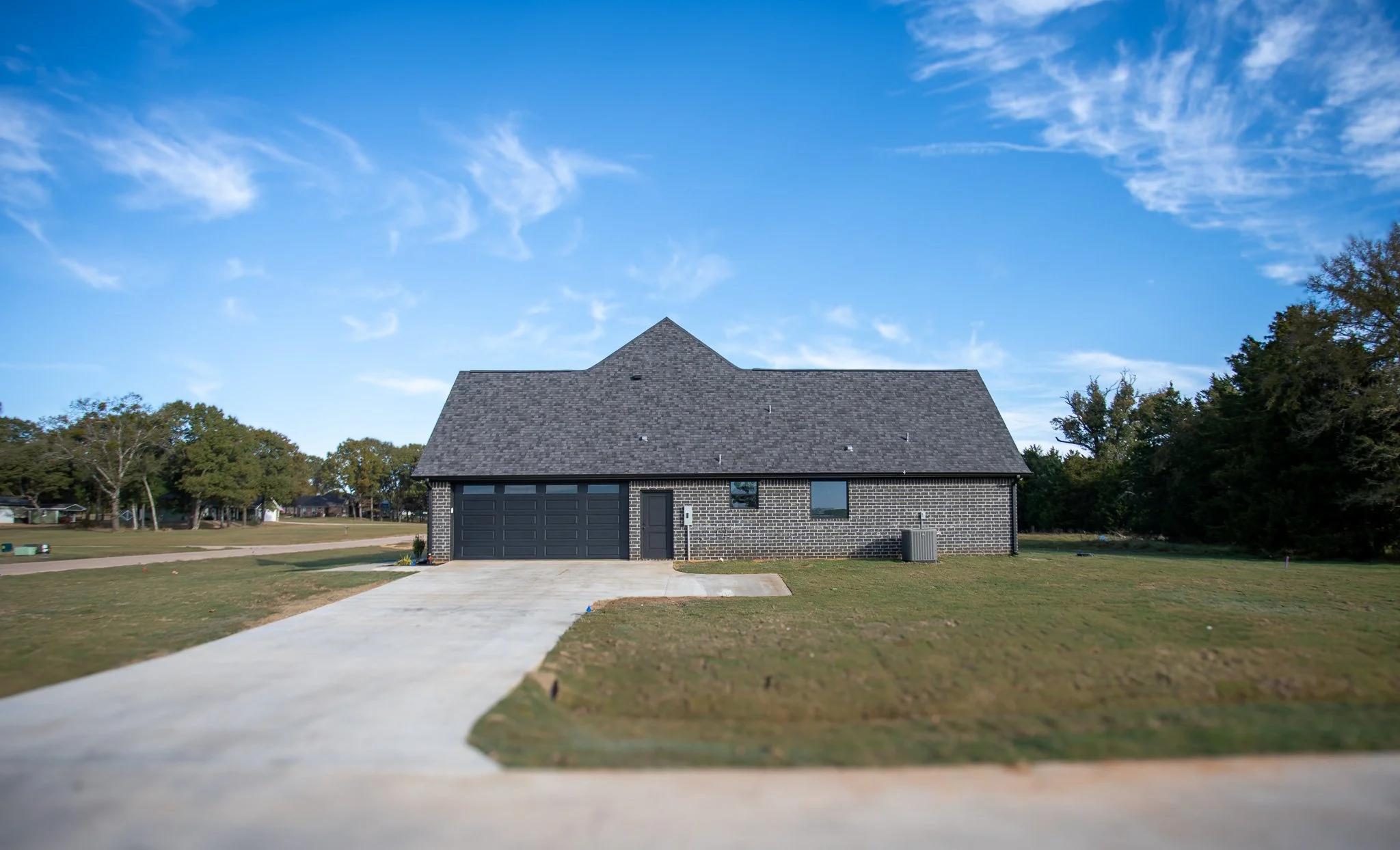 A modern, gray brick house with a dark gray roof and garage door, single front door, and small windows, surrounded by a grassy yard under a bright blue sky with scattered clouds.