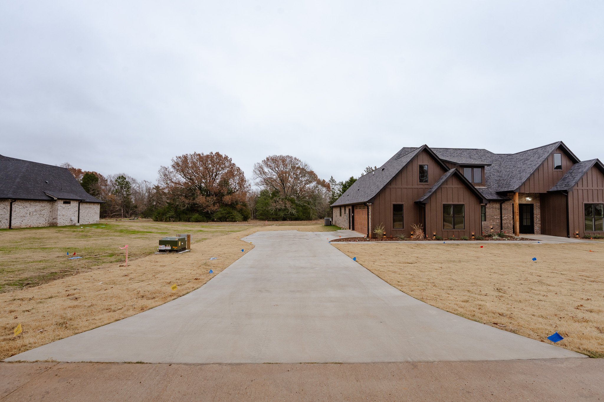 Newly paved concrete driveway leading to a new house with brown siding, a manicured lawn, and several trees in the background.