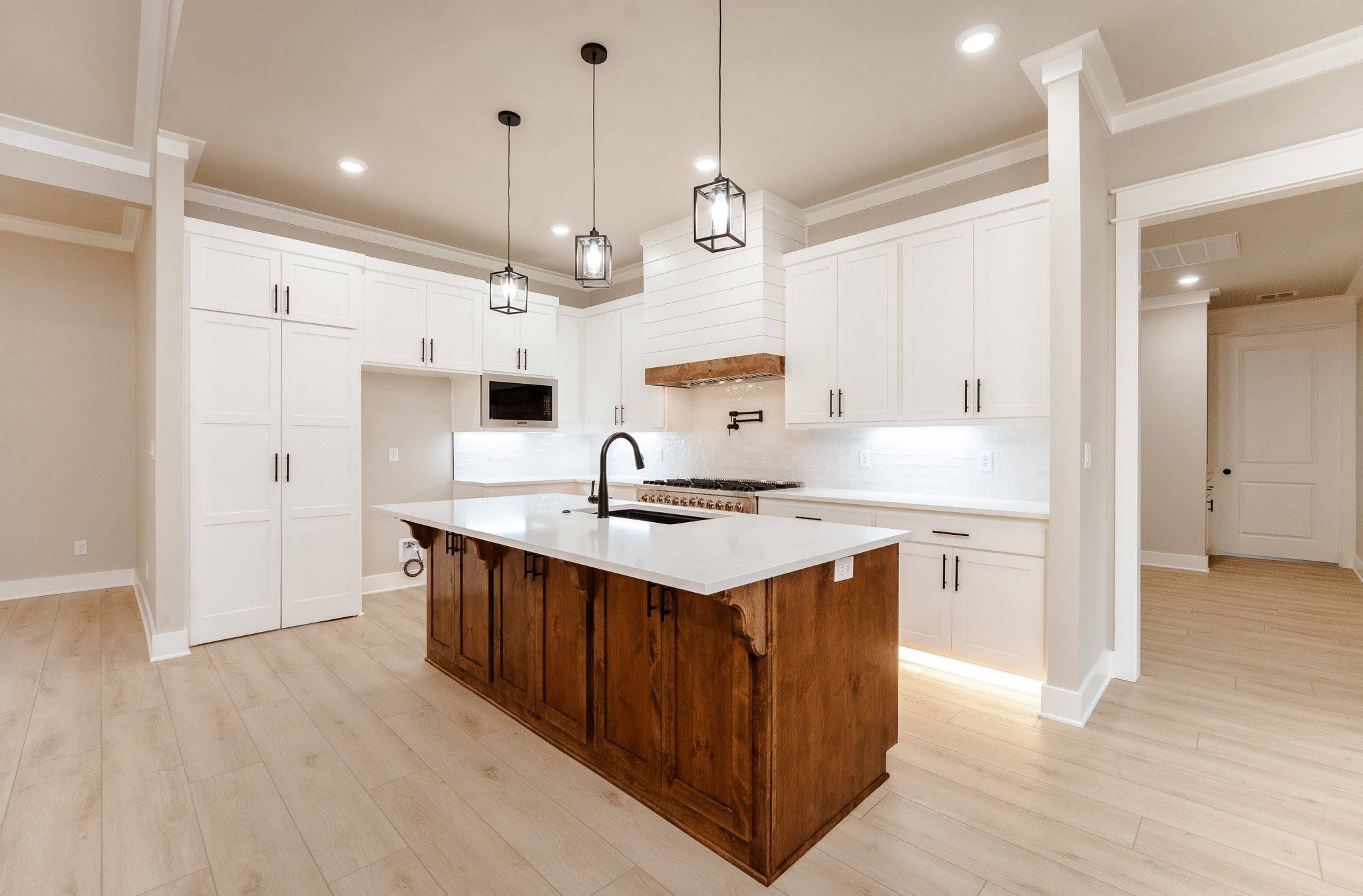 Modern white kitchen with a central island featuring dark wood cabinets and a white countertop, pendant lighting, built-in microwave, and stainless steel stove, with light-colored wood flooring.