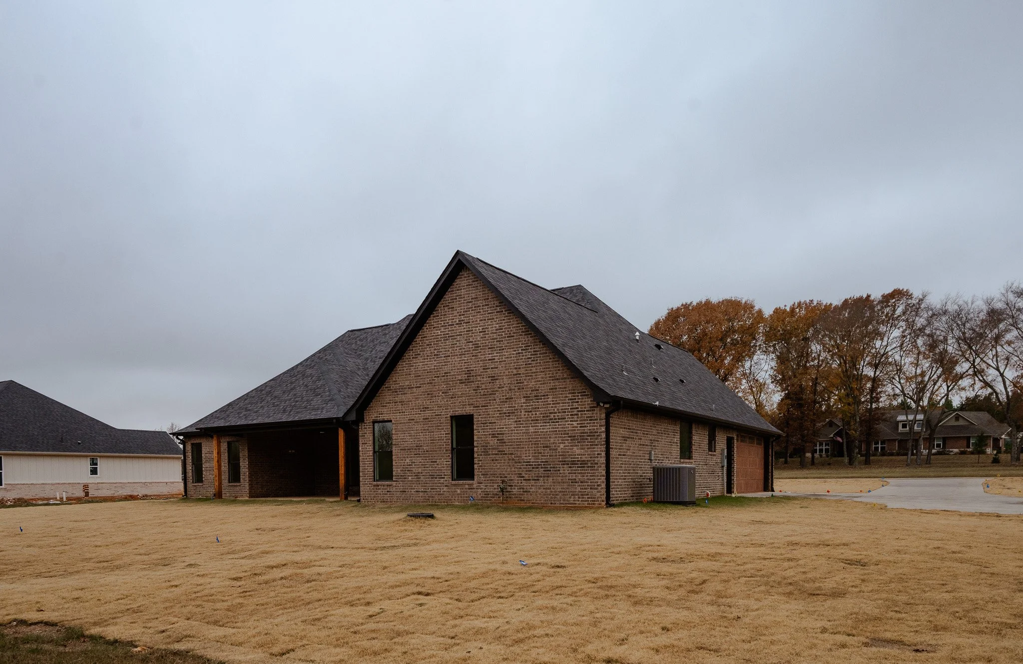 A new brick house under construction with a black roof, surrounded by a grassy yard and neighboring houses in the background on a cloudy day.