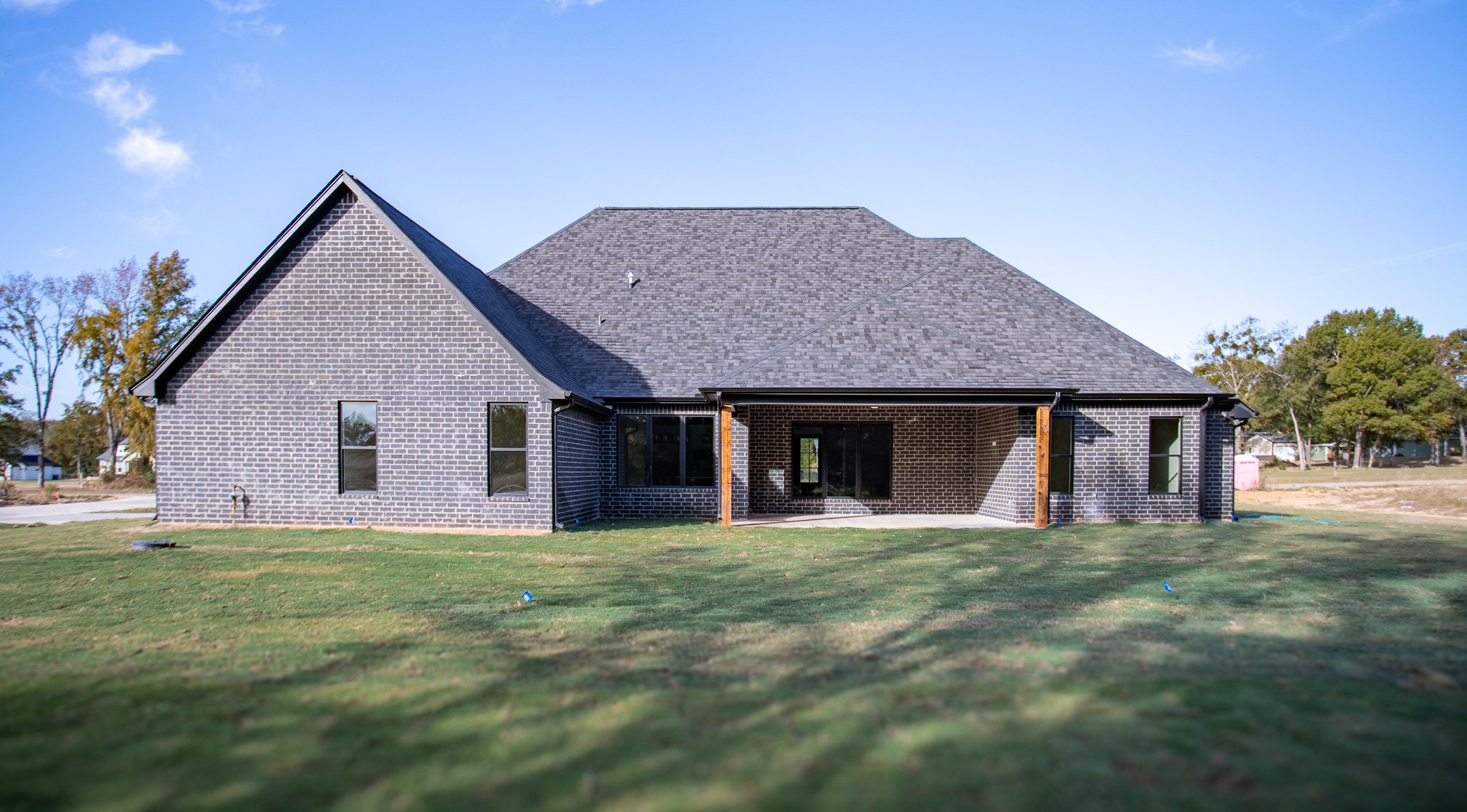 New brick house with a gray shingled roof and a covered back patio, under a blue sky with trees in the background.