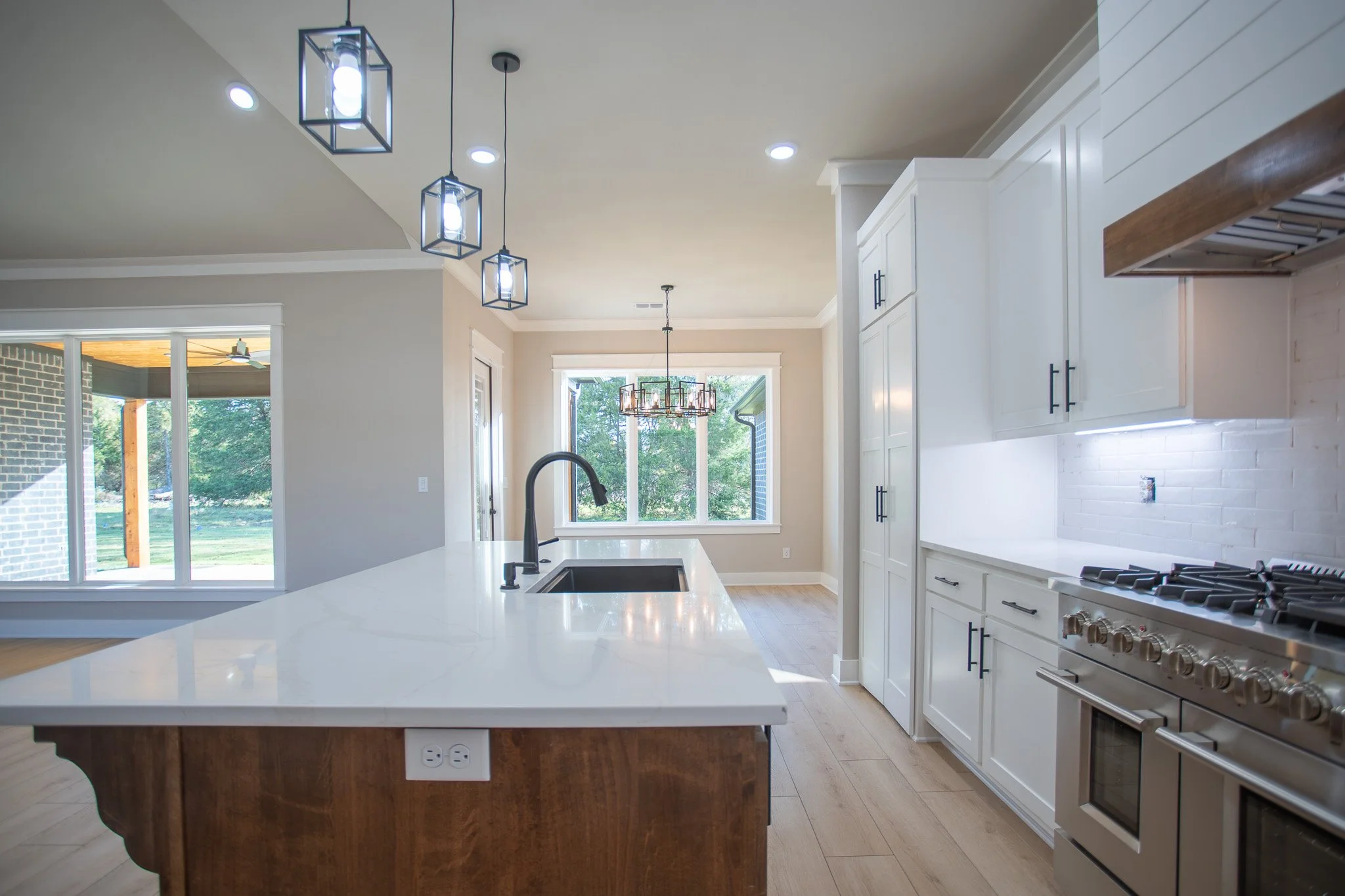 Modern kitchen with white cabinets, stainless steel oven, white countertops, black fixtures, and large windows showing outdoor greenery.
