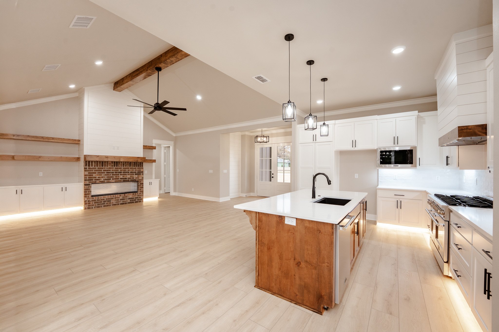 Open-concept kitchen and living area with white cabinets, a kitchen island with a black sink, light wood flooring, and a brick fireplace with wooden shelves, ceiling fans, and pendant lighting.