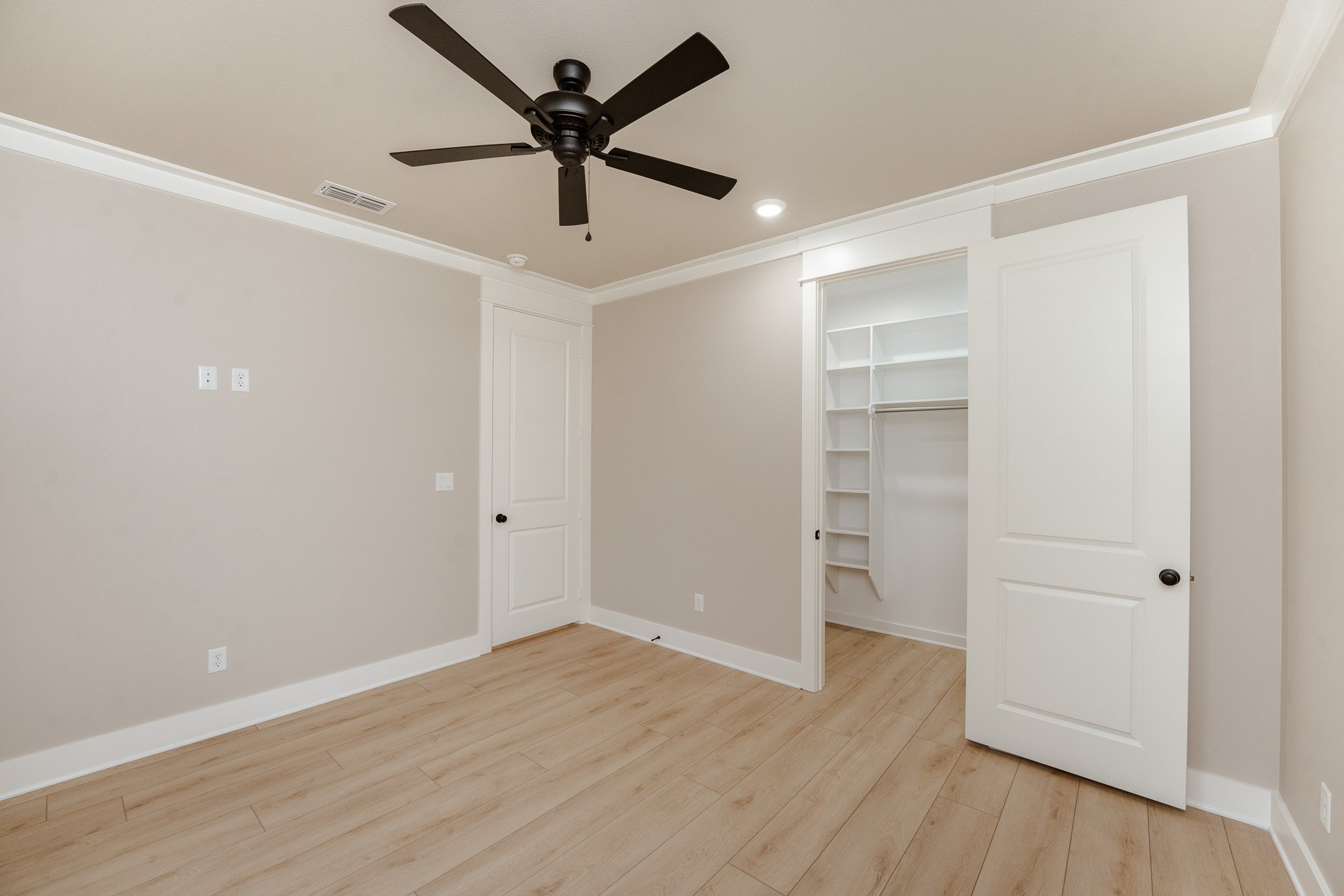 Empty bedroom with beige walls, wood flooring, a ceiling fan, two white doors, and a walk-in closet with built-in shelves.