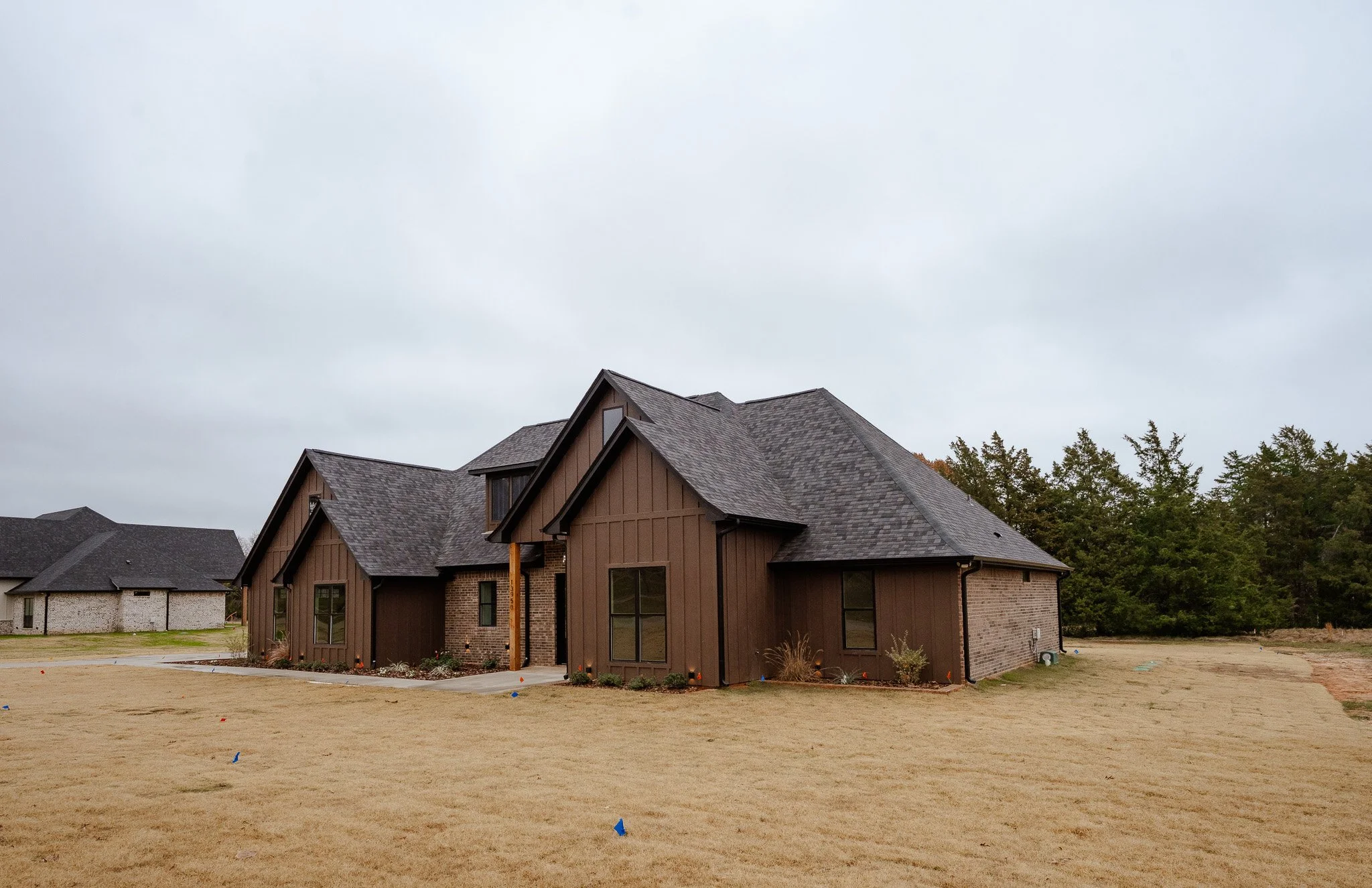 A new two-story house with brown siding and a shingled roof, situated on a grassy lot with some small plants near the foundation and neighboring houses in the background under an overcast sky.