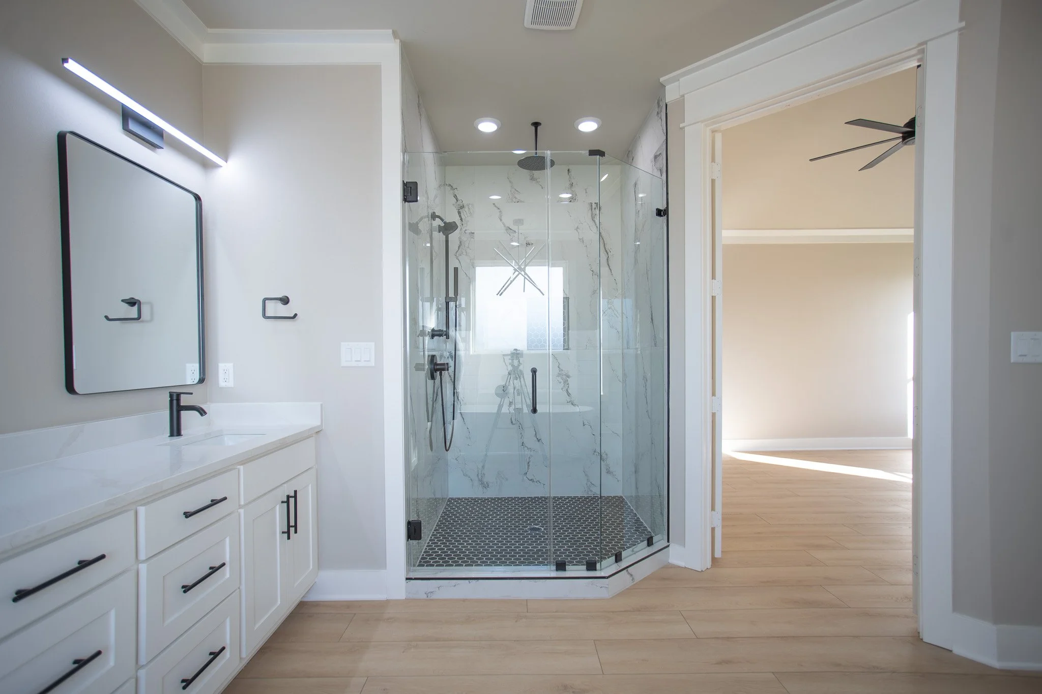 Modern bathroom with double vanity, black hardware, large mirror, and glass-enclosed walk-in shower with marble tiles.