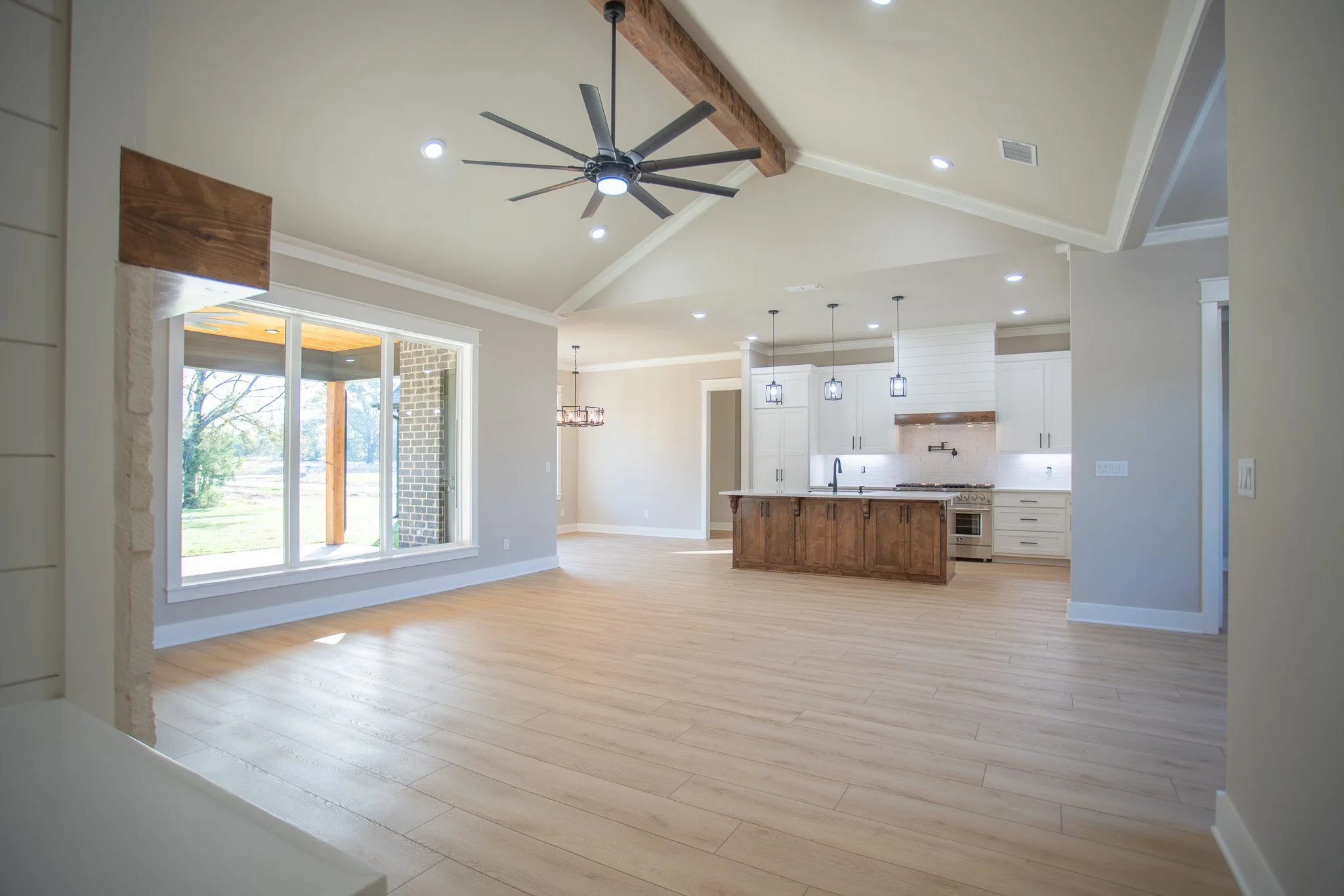 Open-concept kitchen and living area with light wood floors, white walls, and large window showing outdoor scenery. The kitchen has white cabinets, a wooden island with a dark countertop, and modern lighting fixtures. Ceiling with recessed lighting a