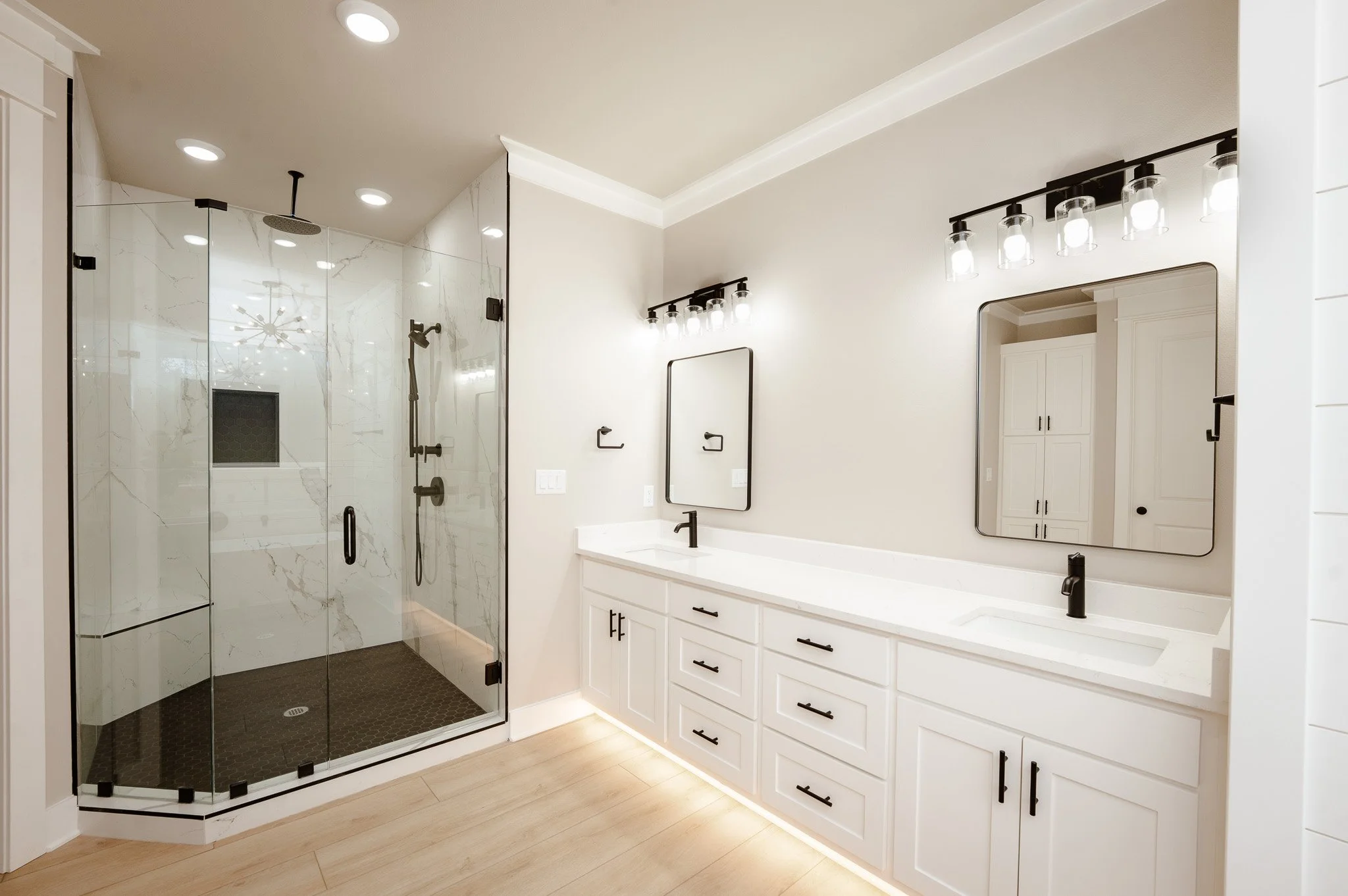 Modern bathroom with glass enclosed shower, double vanity with two mirrors and black fixtures, light wood flooring, and white cabinetry.