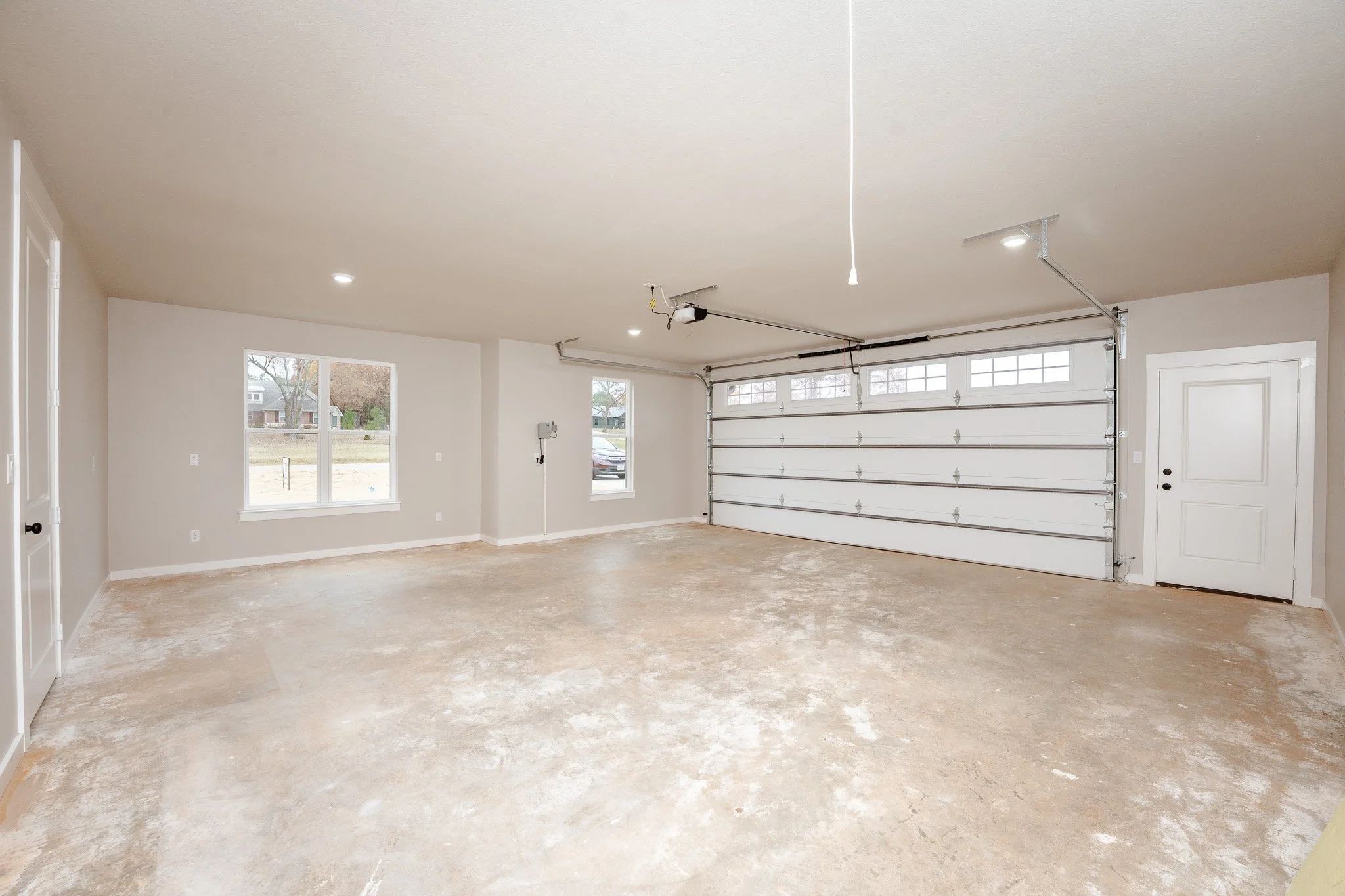 Empty residential garage with a concrete floor, white walls, large window, and a white sectional garage door. There is a small door on the right side, and a wall-mounted utility box on the wall near the window.