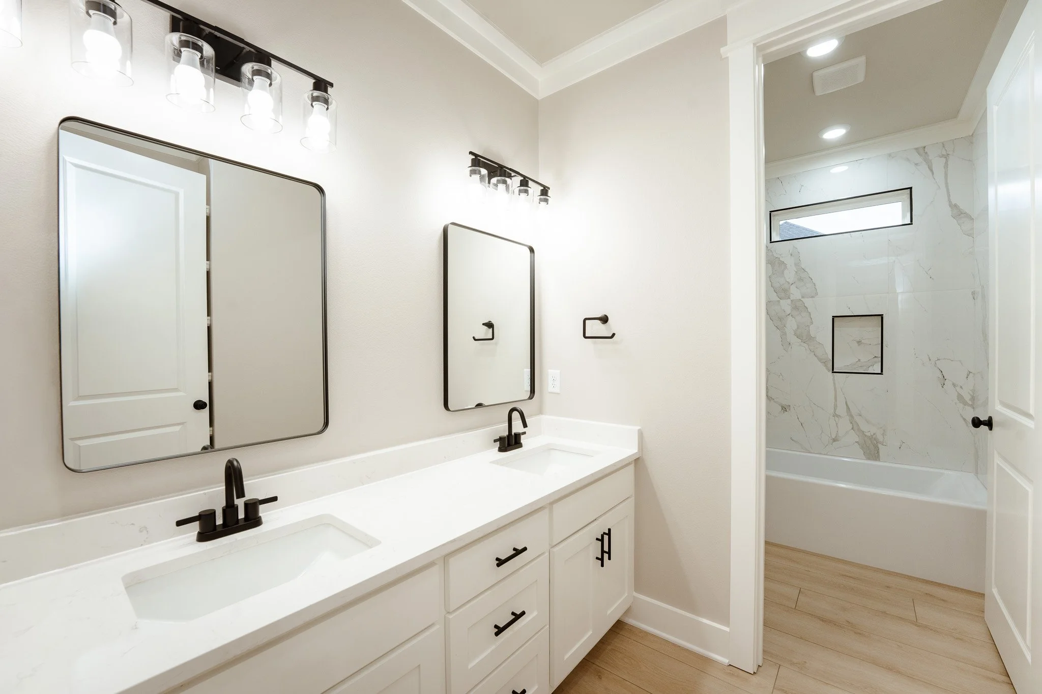 Modern bathroom with dual sinks, large mirrors, and black fixtures. Light wood flooring and a separate shower area with white marble walls and a small rectangular window.