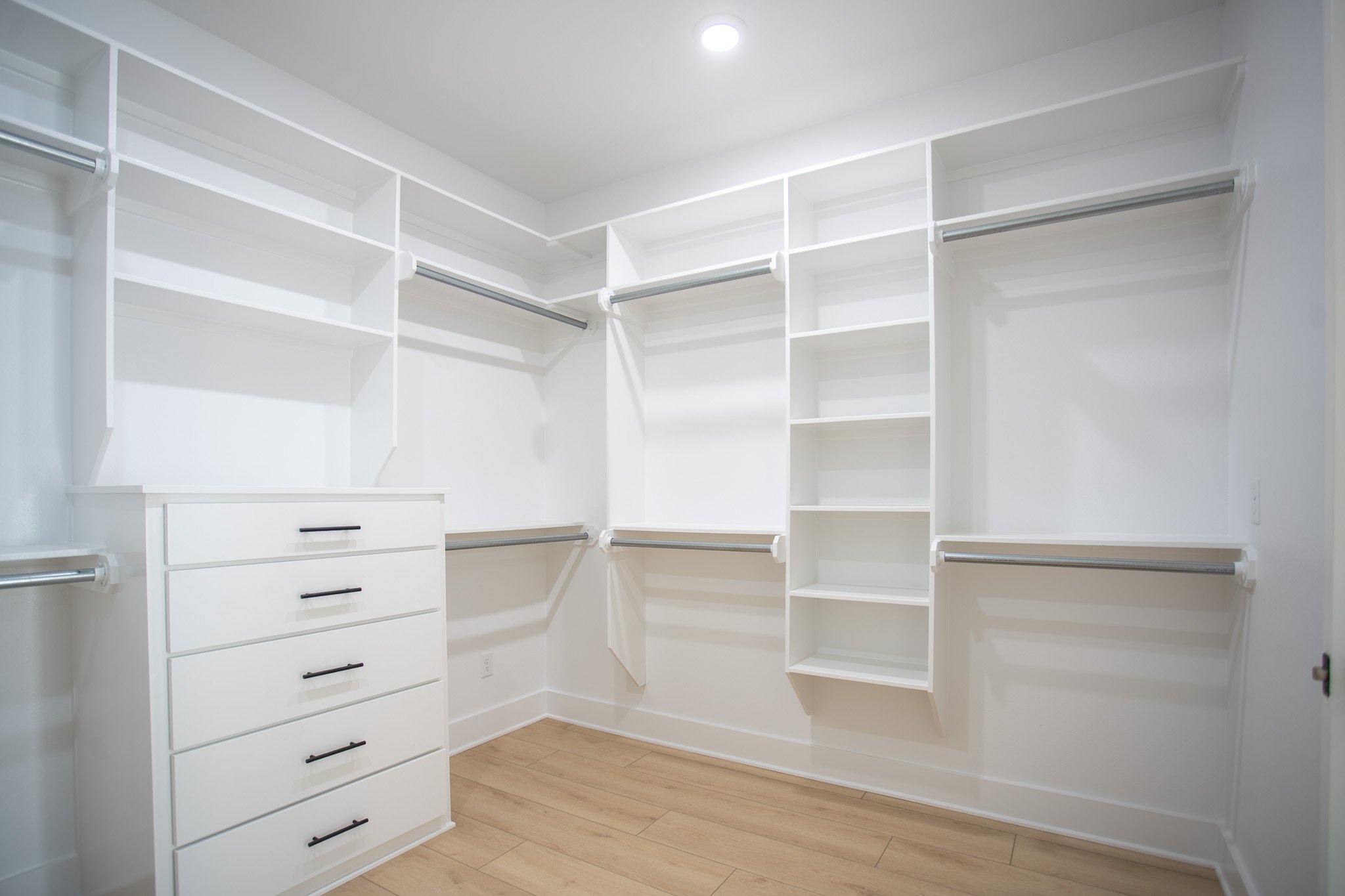 Empty white walk-in closet with built-in shelves and hanging rods, beige hardwood floors, and recessed ceiling light.