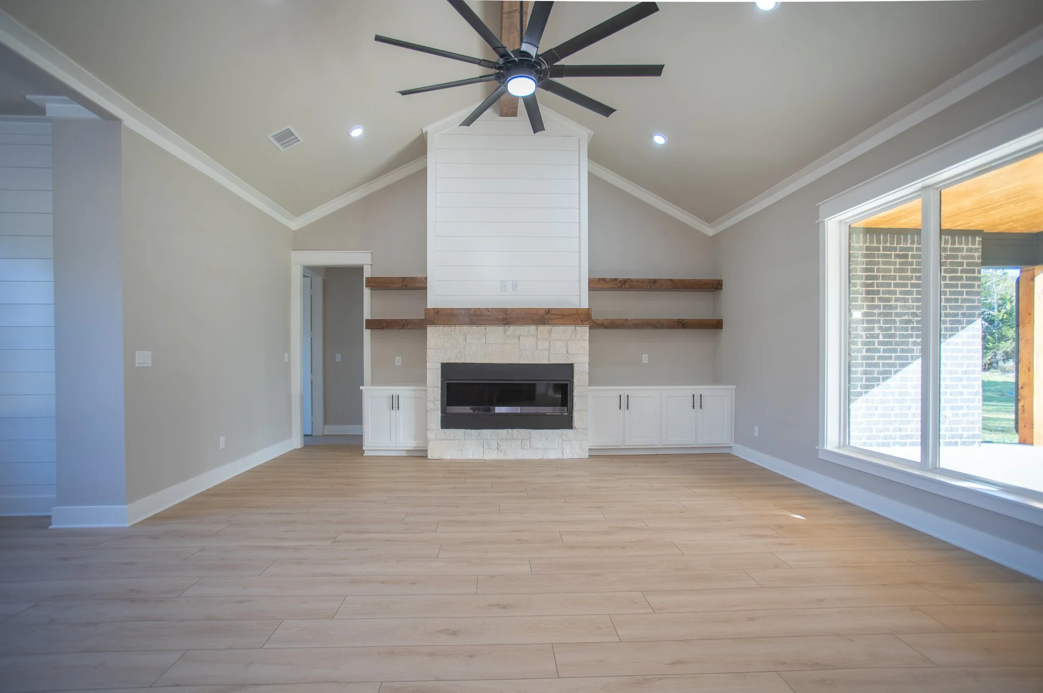 Empty living room with light wood flooring, a stone fireplace with built-in white cabinets, wooden shelves, a large window, a ceiling fan, and recessed lighting.